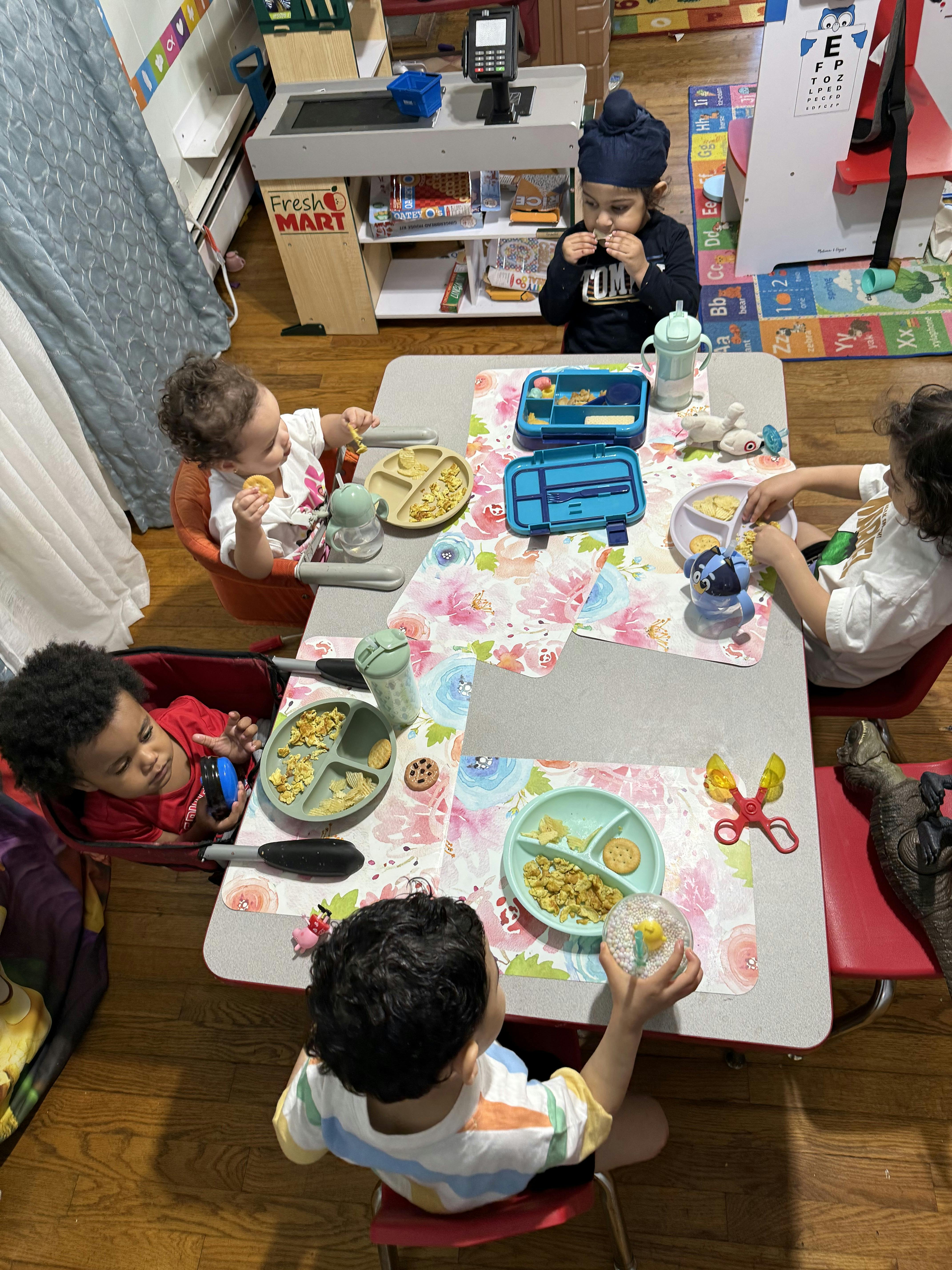 Five children eating snacks and playing at a table in a playroom