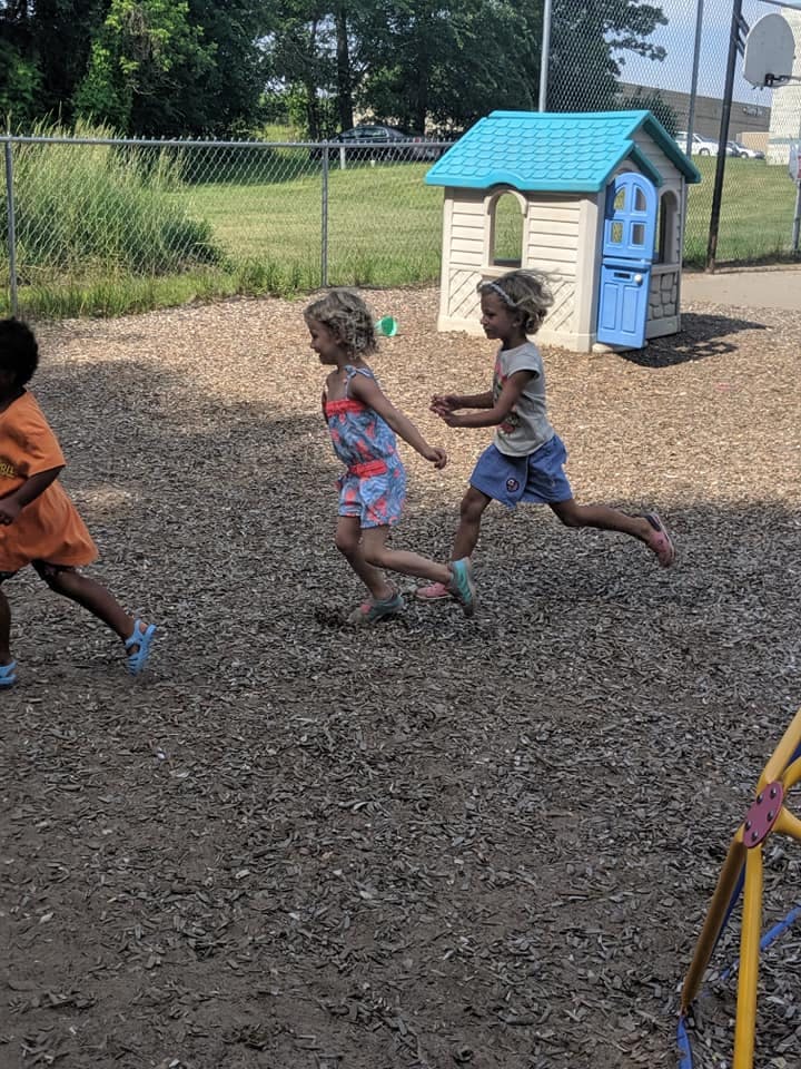 Three children running on wood chips near a playhouse
