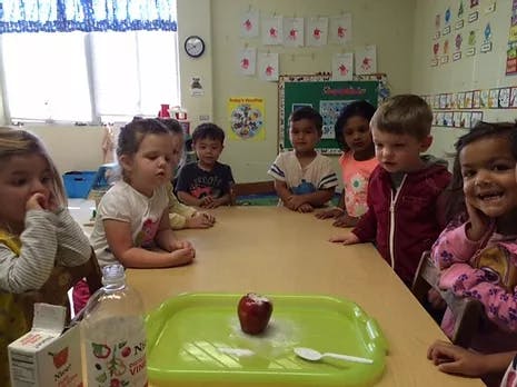 Children gathered around a table with an apple on a tray in a classroom