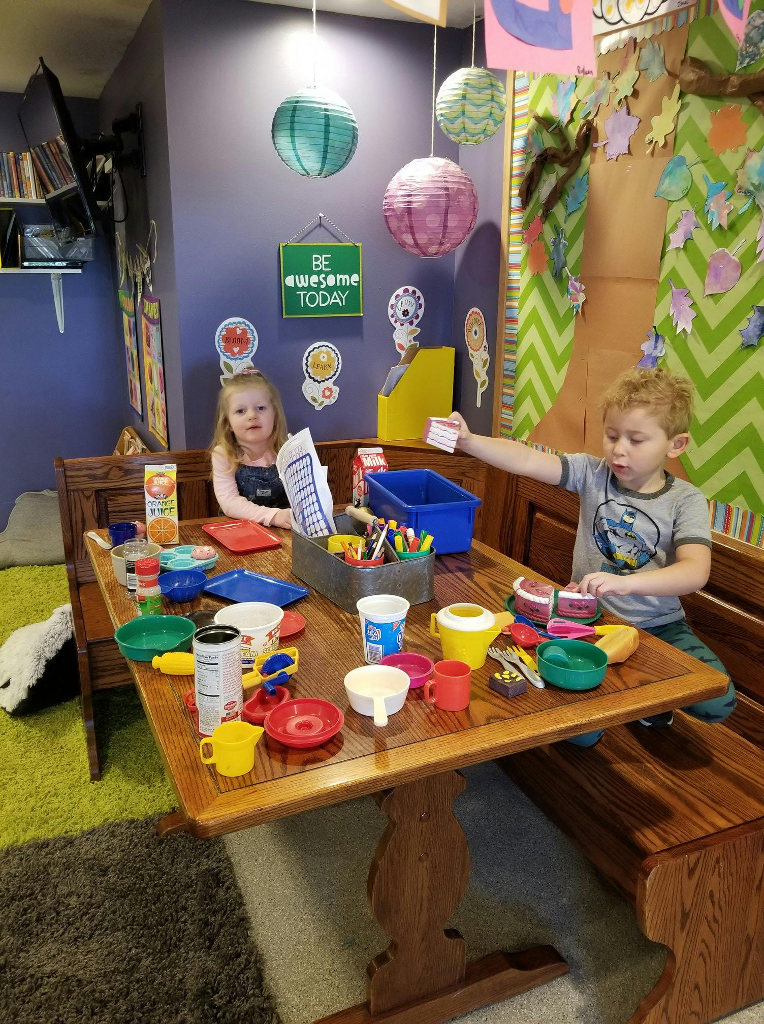 Two children playing with toy dishes and art supplies at a wooden table