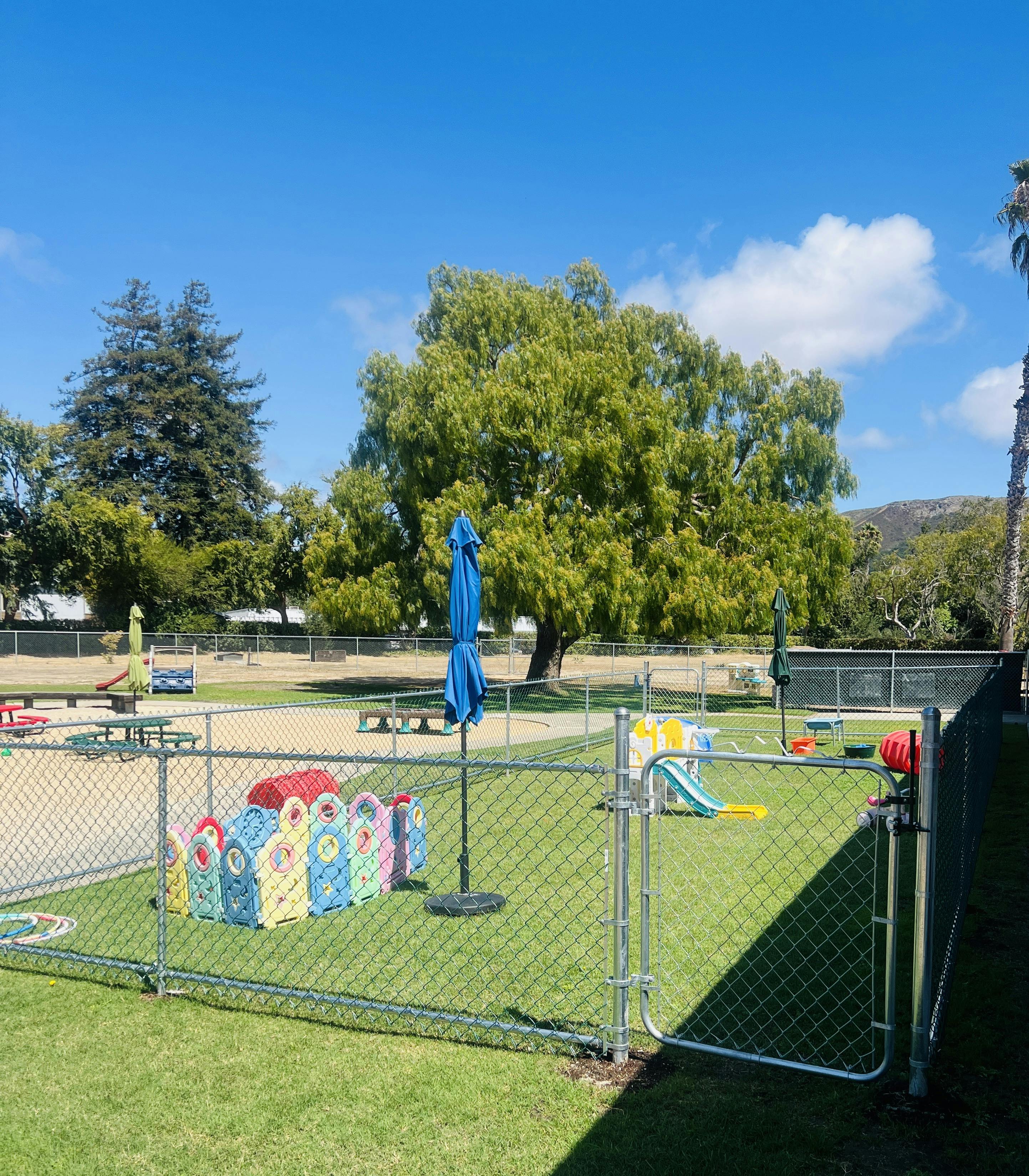 Fenced outdoor play area with a blue umbrella and colorful play structures
