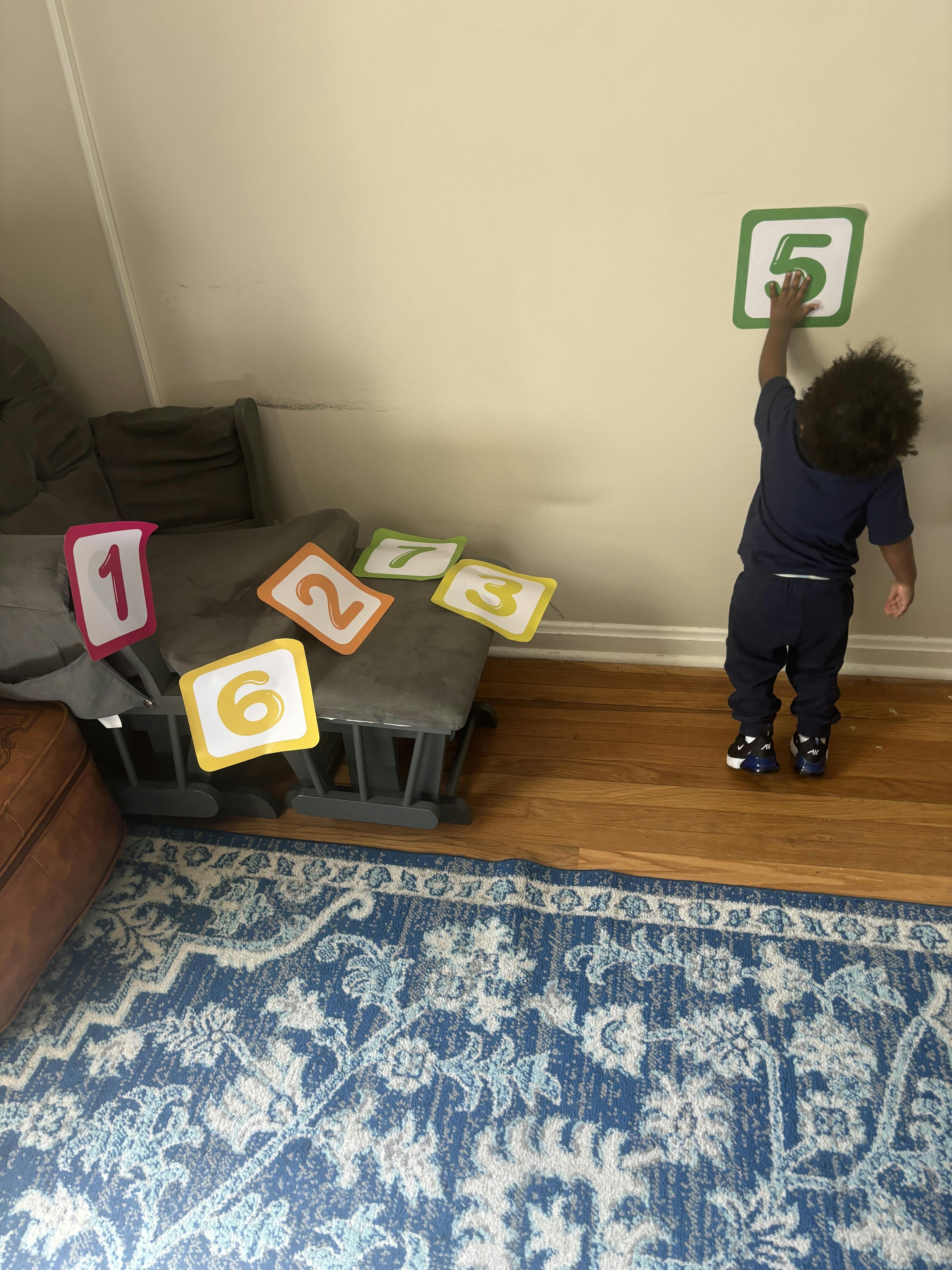 Child placing a number 5 on a wall, with other numbers on a chair