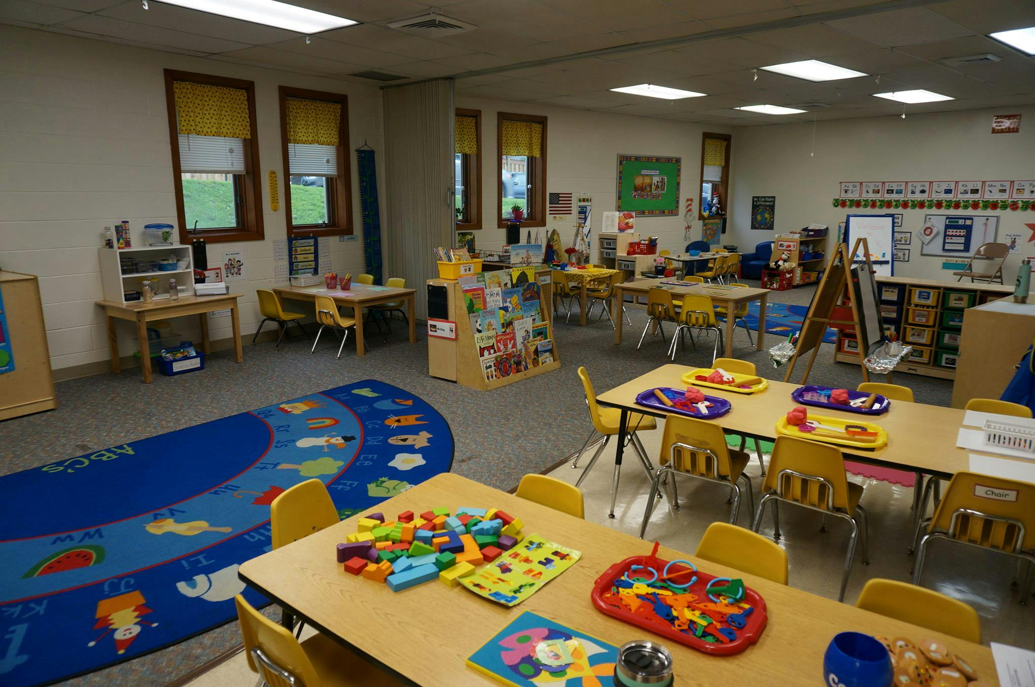 Empty classroom with small tables and chairs, a blue rug, and toys on tables