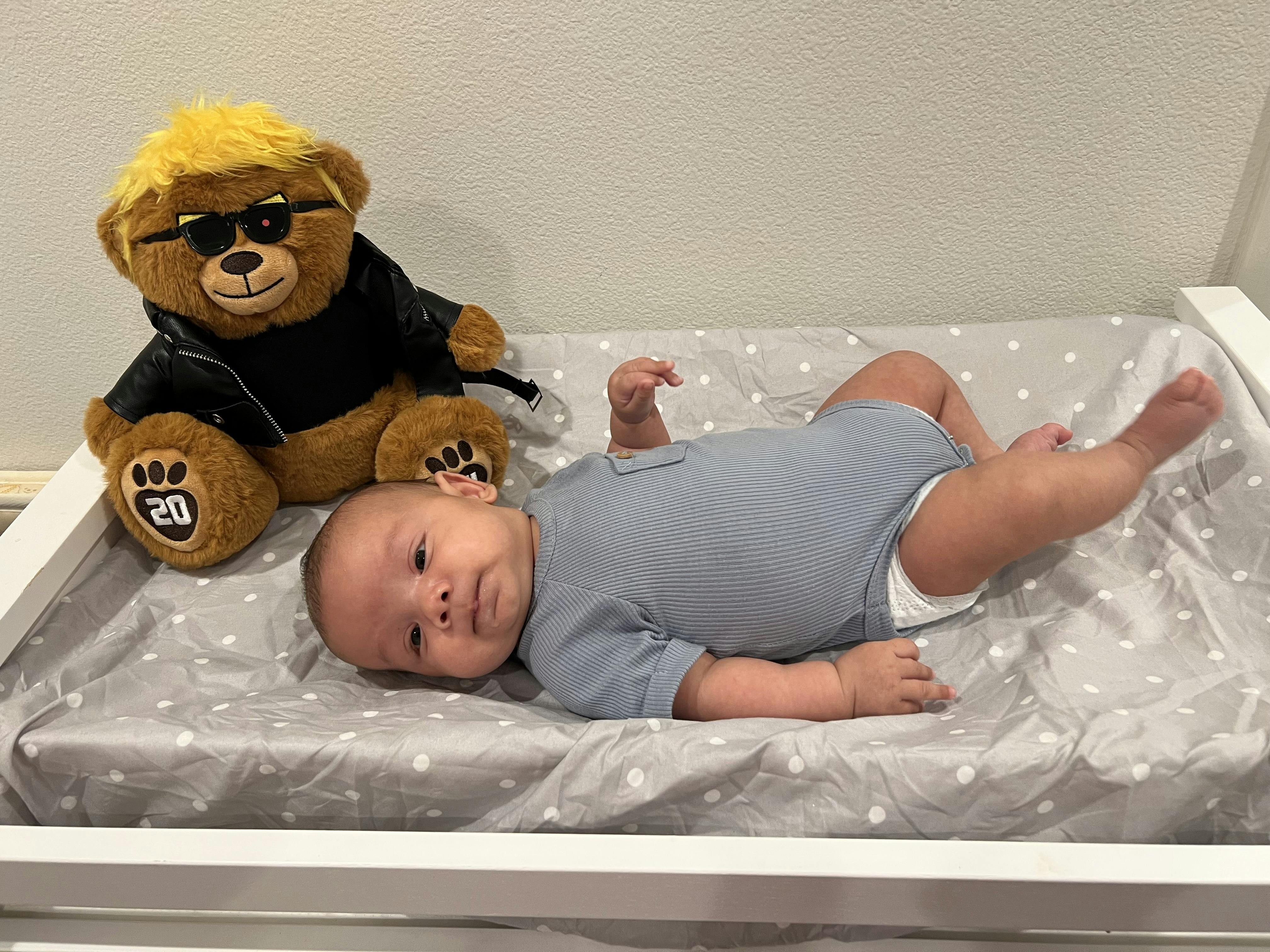Child in blue onesie on a polka dot changing pad next to a teddy bear