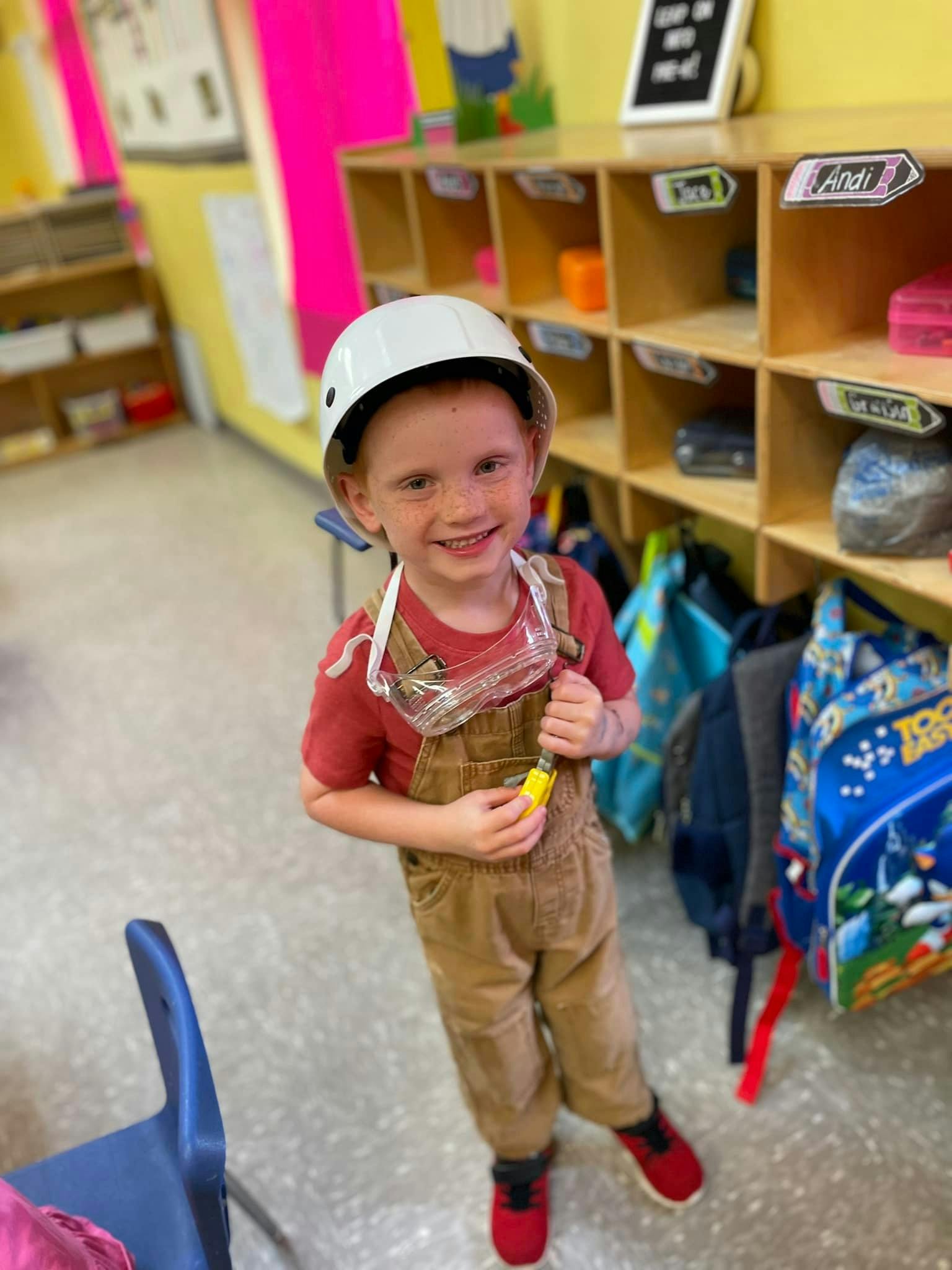 Child wearing a white helmet, safety goggles, and overalls in a classroom