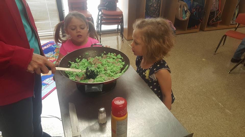Person stirring green food in a pan with two children watching