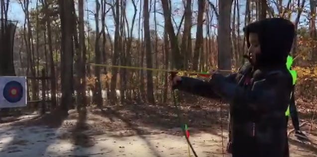Child aiming a bow and arrow at a target in a wooded area