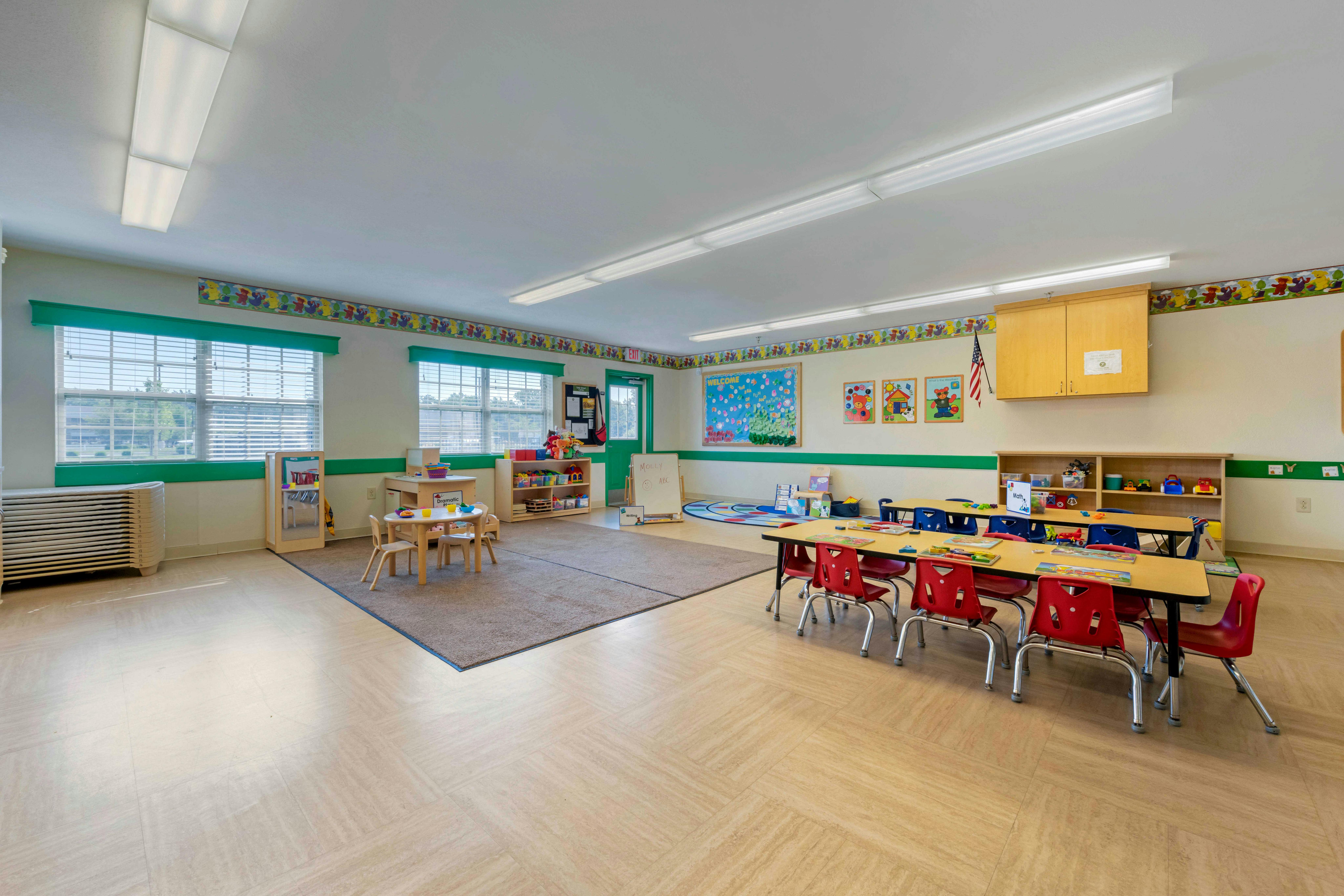 A classroom with child-sized tables and chairs, a rug, and shelves with toys