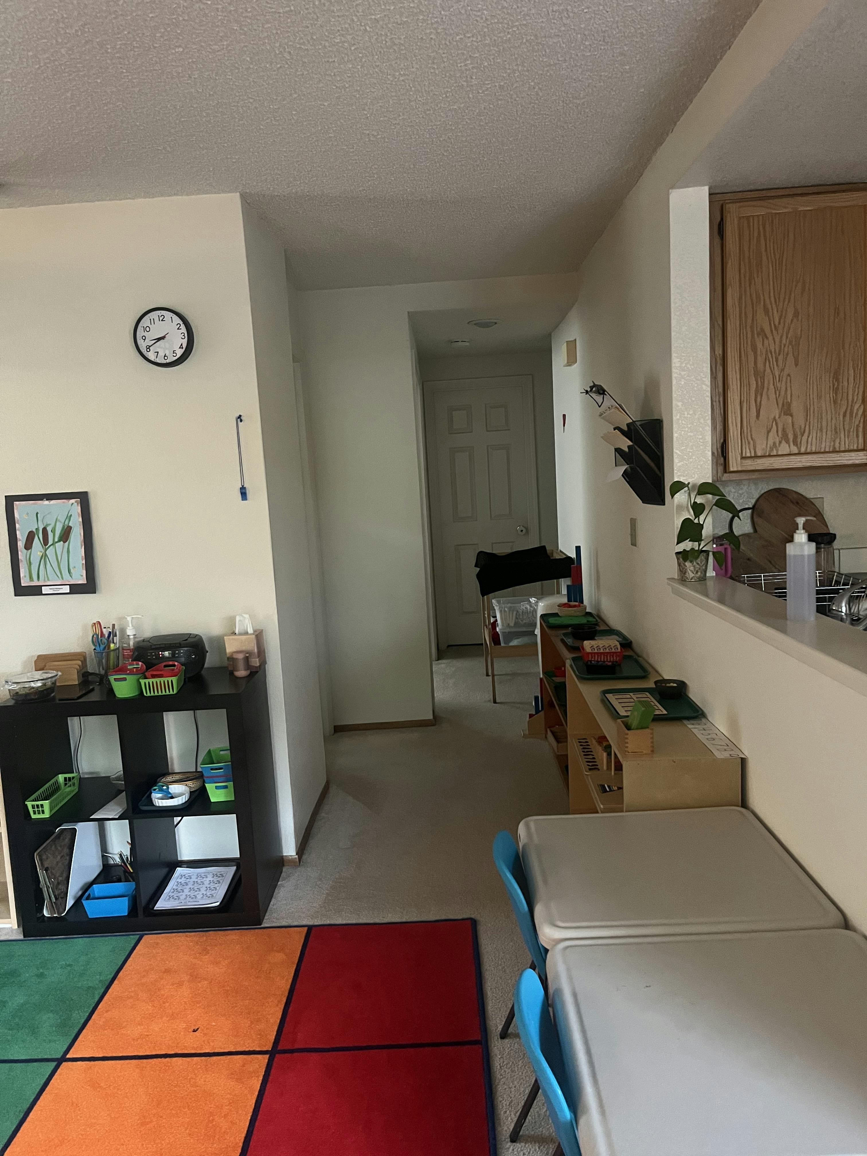 Classroom interior with small tables and chairs, shelves of learning materials, and colorful rug