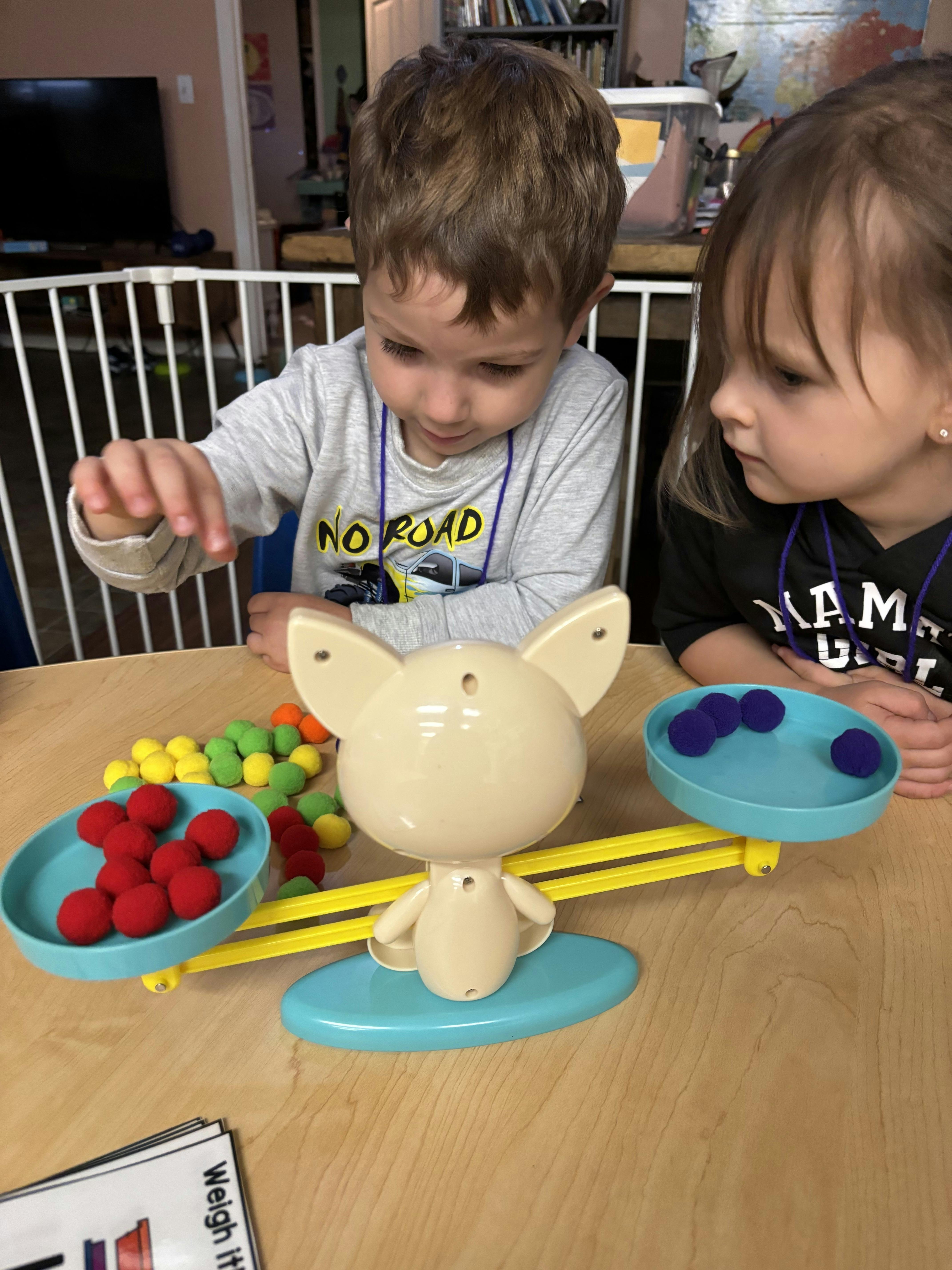 Two children playing with a balance scale and colorful pom-poms