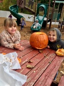 Two children at a picnic table with a carved pumpkin, playground in background