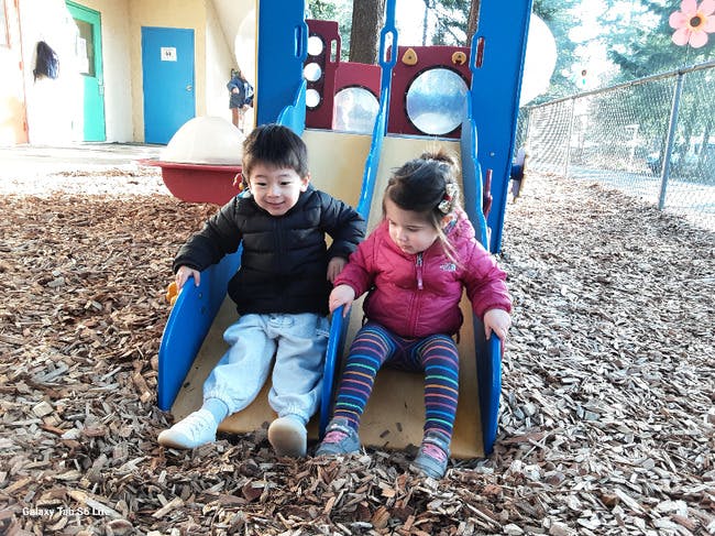 Two children sitting side-by-side at the bottom of a slide on wood chips