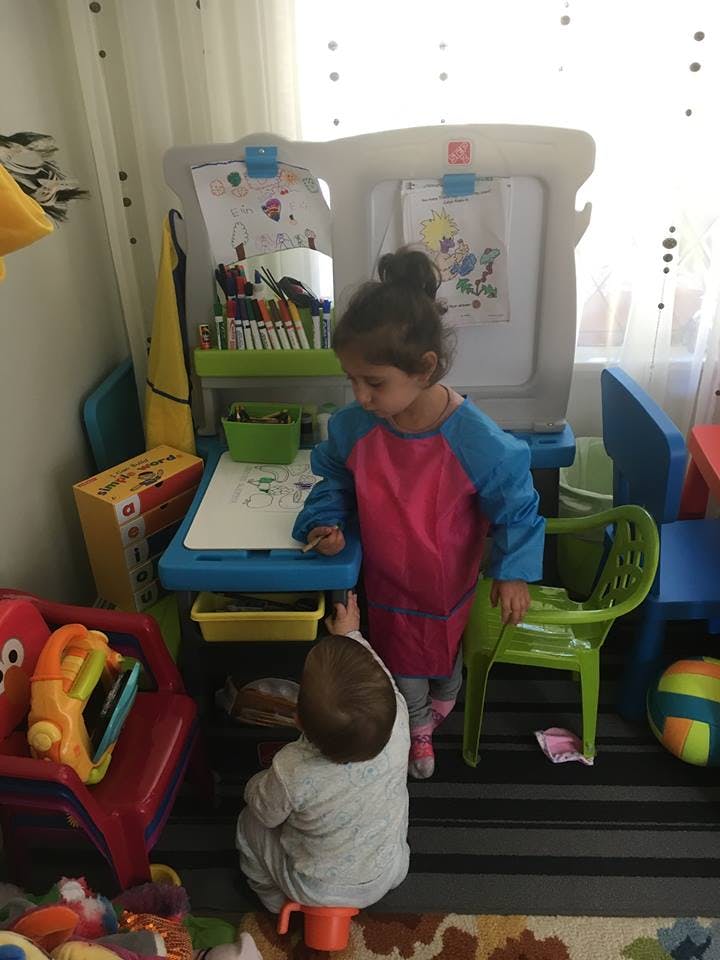Child standing at an art easel with a baby on the floor reaching up