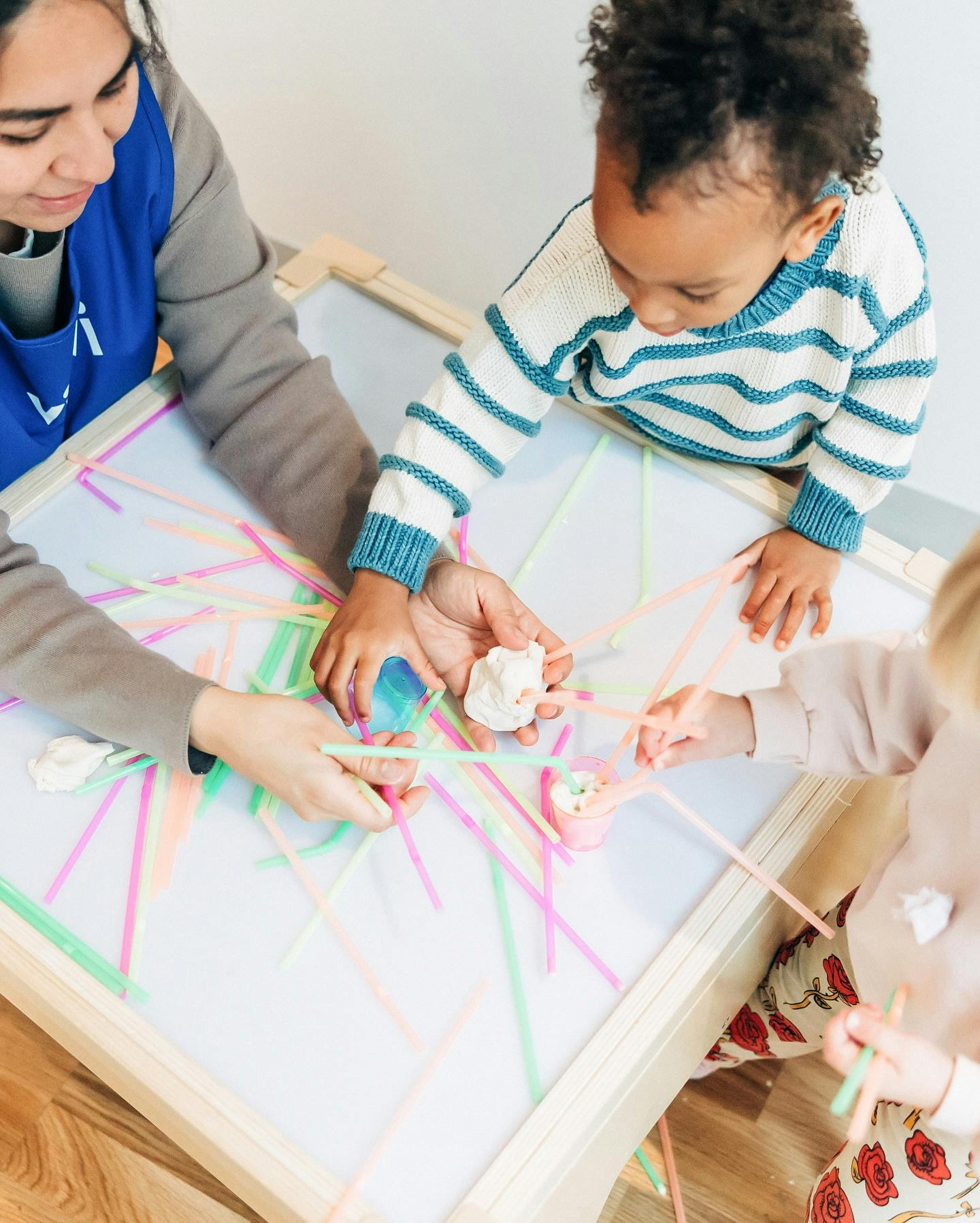 Person and two children playing with straws and clay on a light table