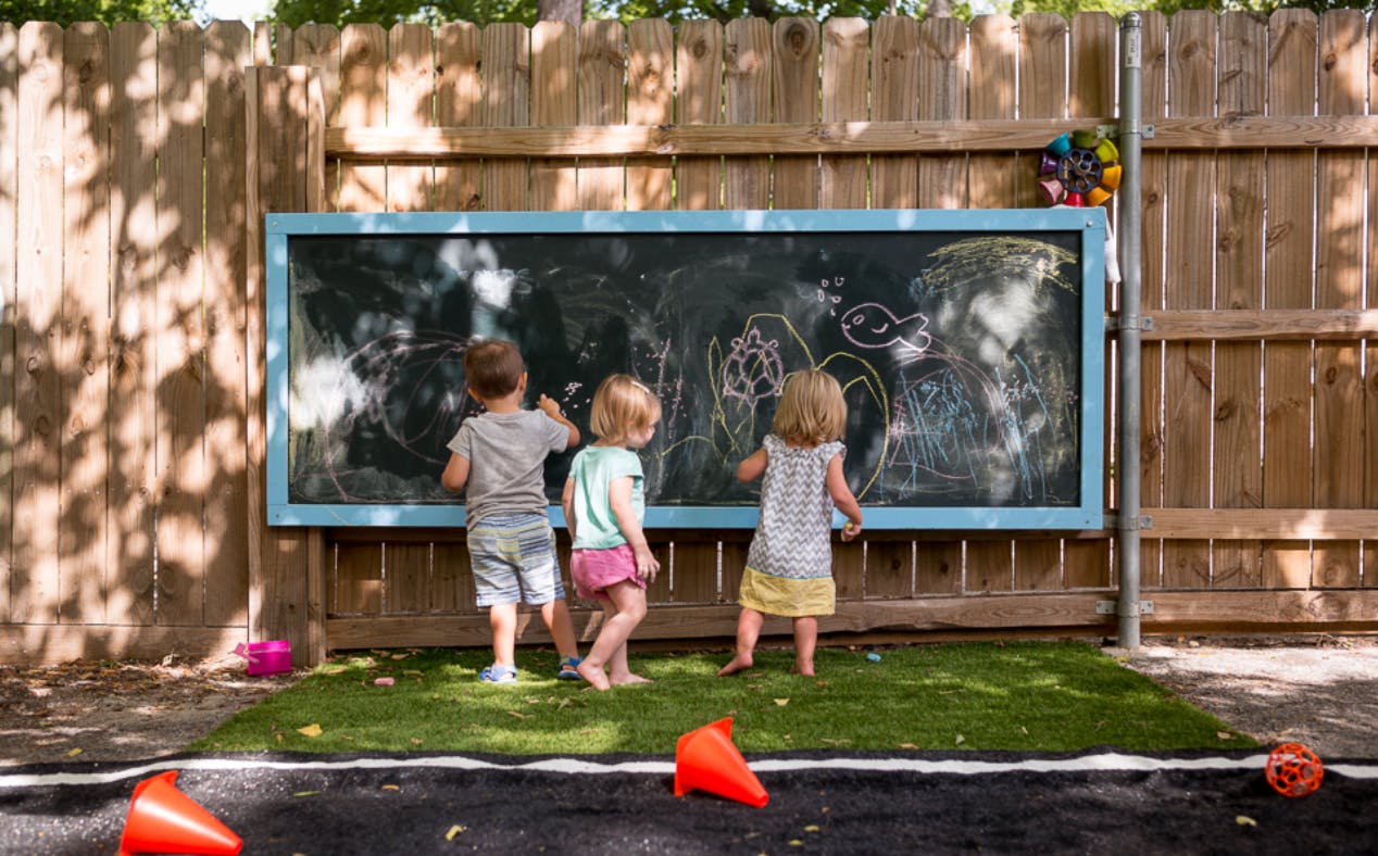 Three children drawing on a large outdoor chalkboard