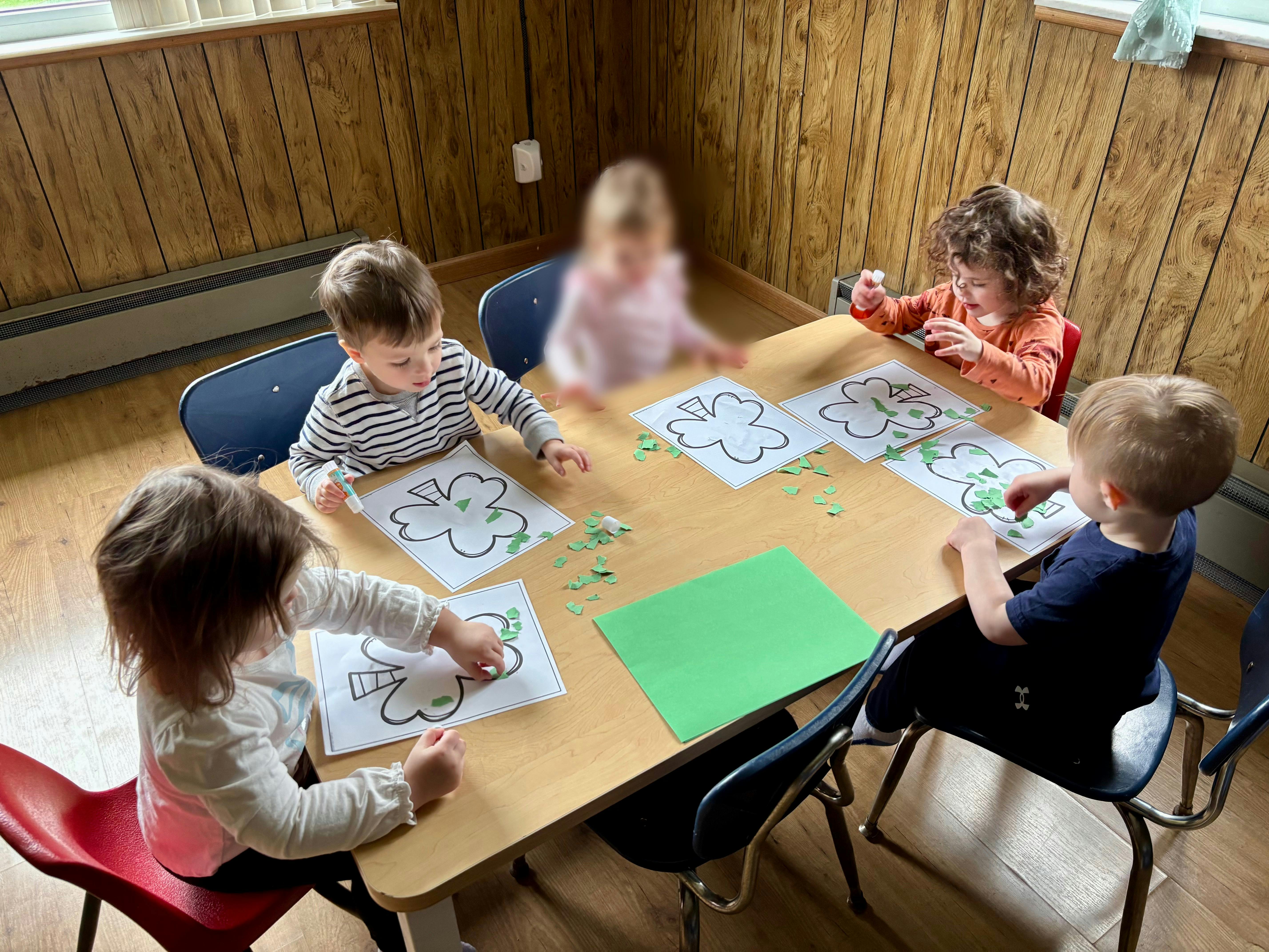 Children gluing green paper scraps onto shamrock outlines at a table