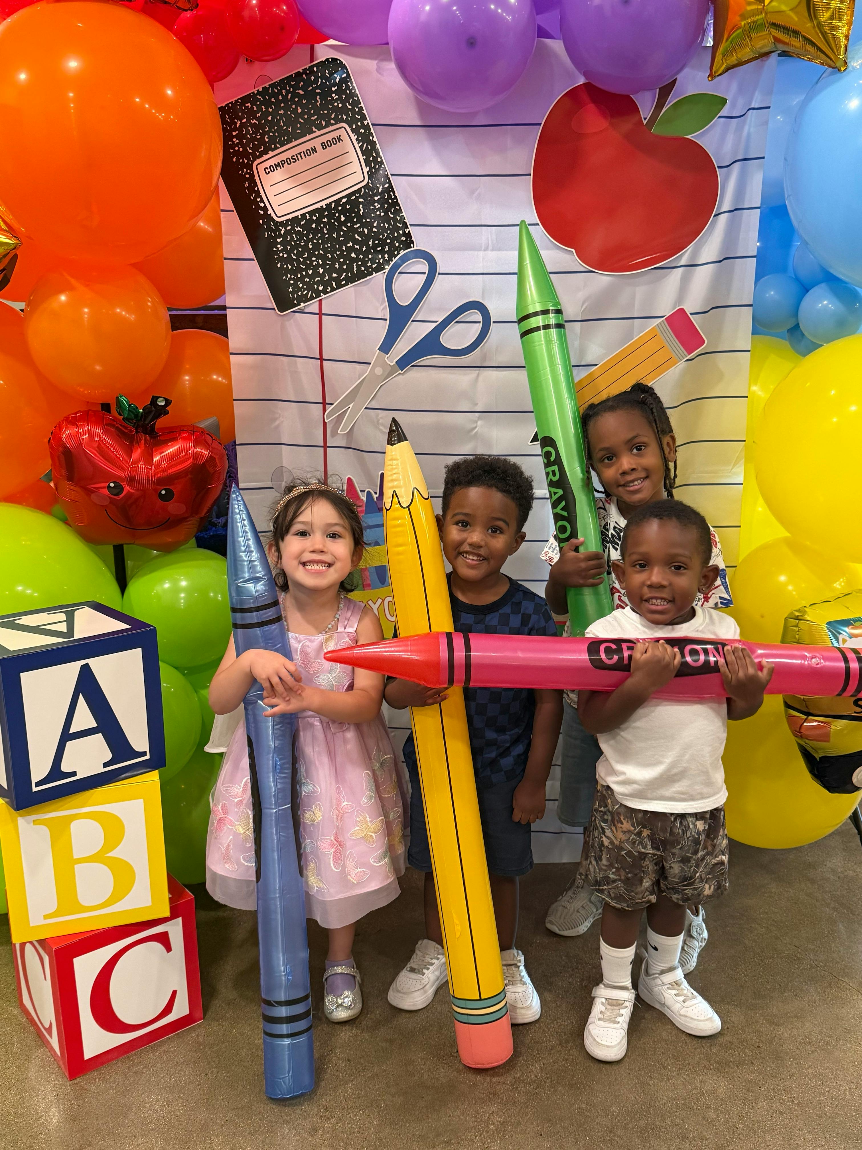Four children holding large inflatable school supply props in front of balloons