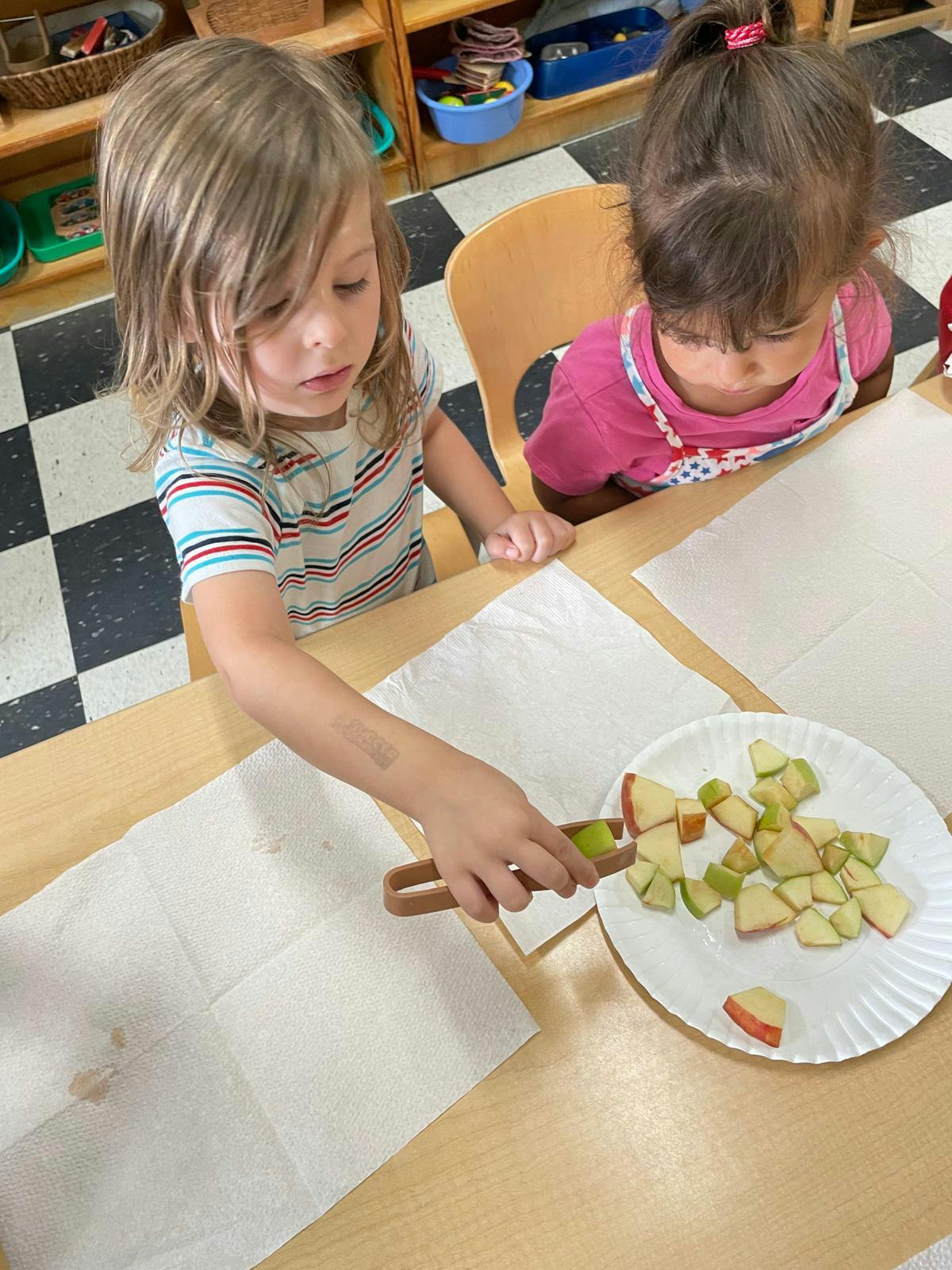 Two children at a table, one using tongs to pick up apple pieces from a plate