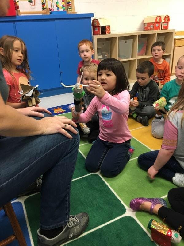 Children sitting on a rug, one child holding a craft project
