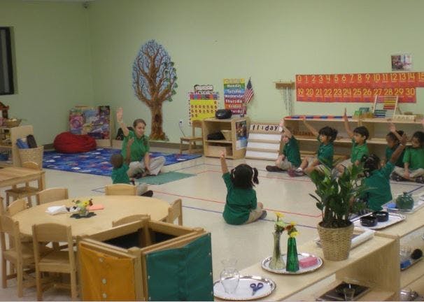 Adult and children sitting on the floor with hands raised in a classroom