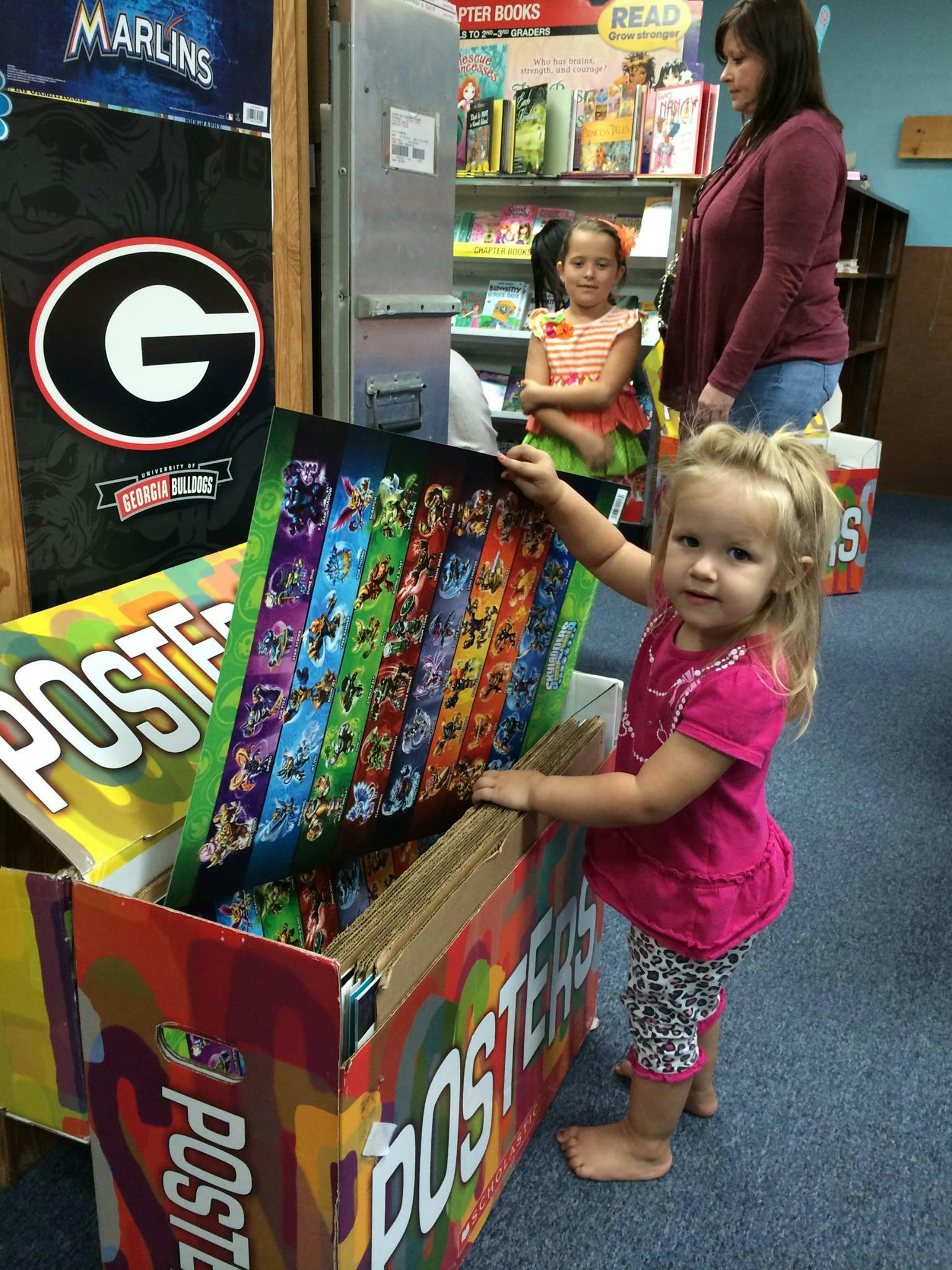 Child looking at posters in a box, with a person and child in the background