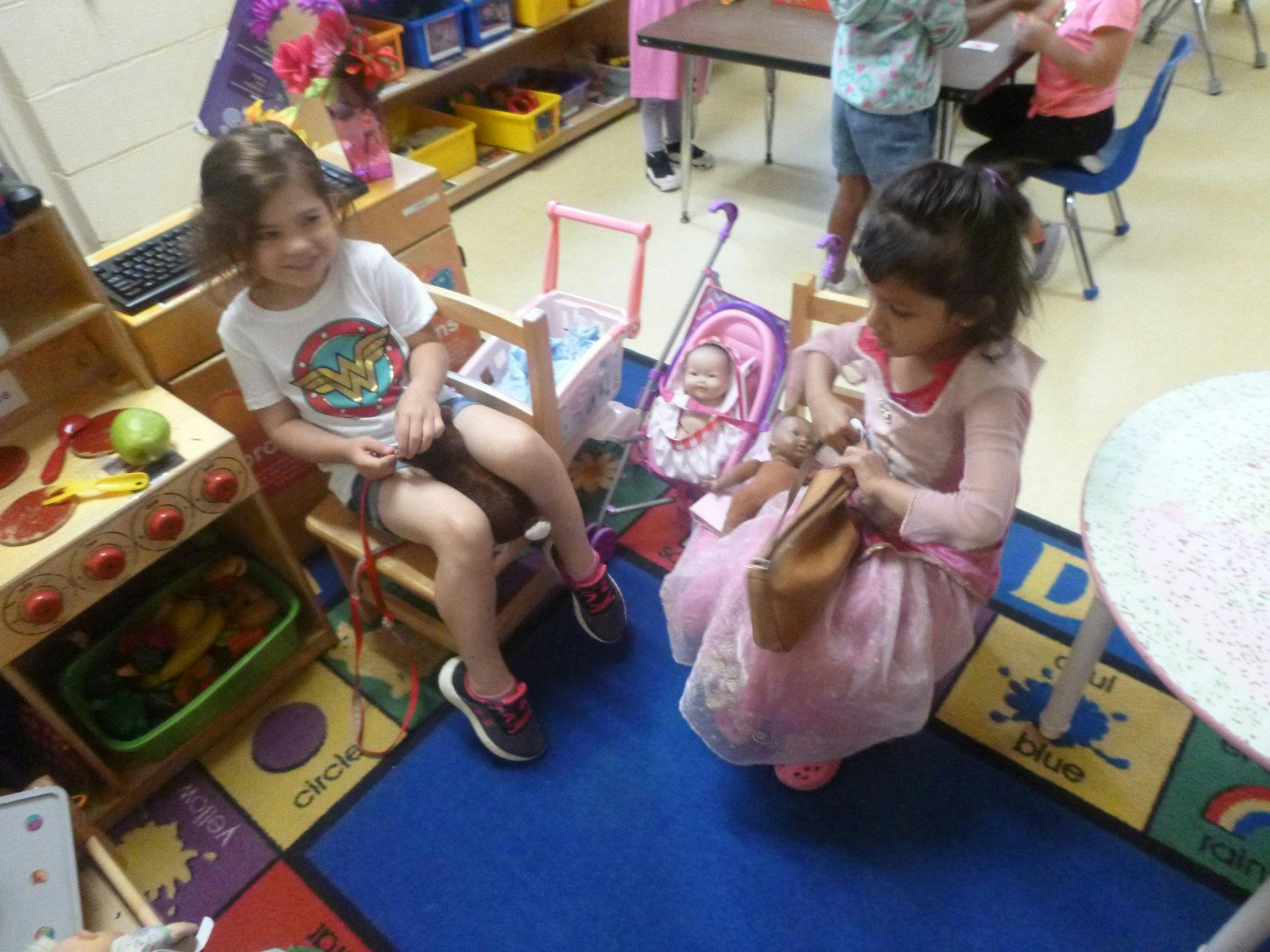 Two children playing with dolls and toy furniture in a classroom