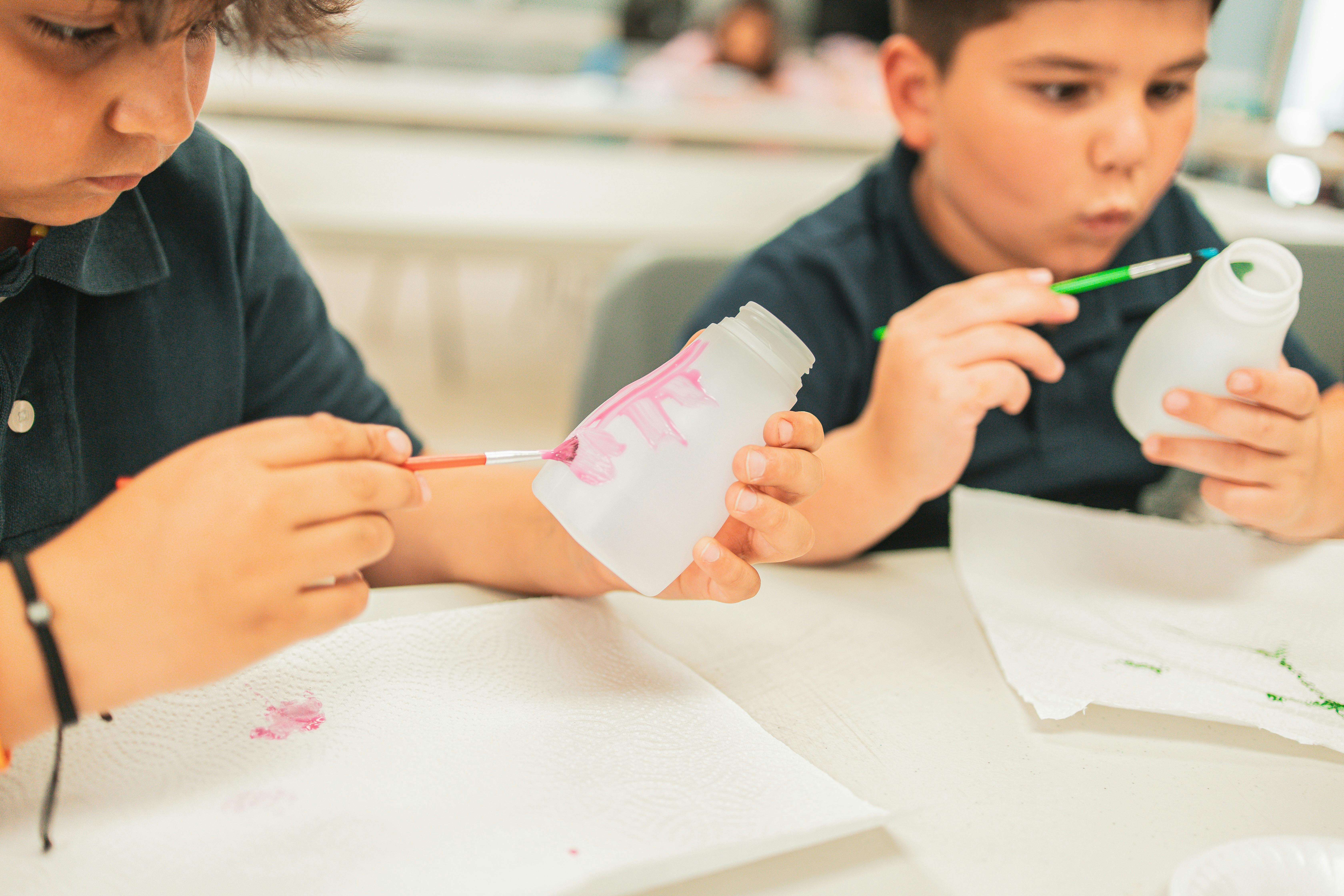 Two children painting small plastic bottles in a classroom