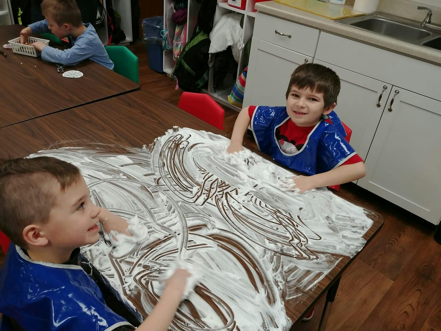Two children playing with shaving cream on a table in a classroom