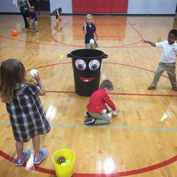 Children playing a game with a decorated trash can on a gym floor