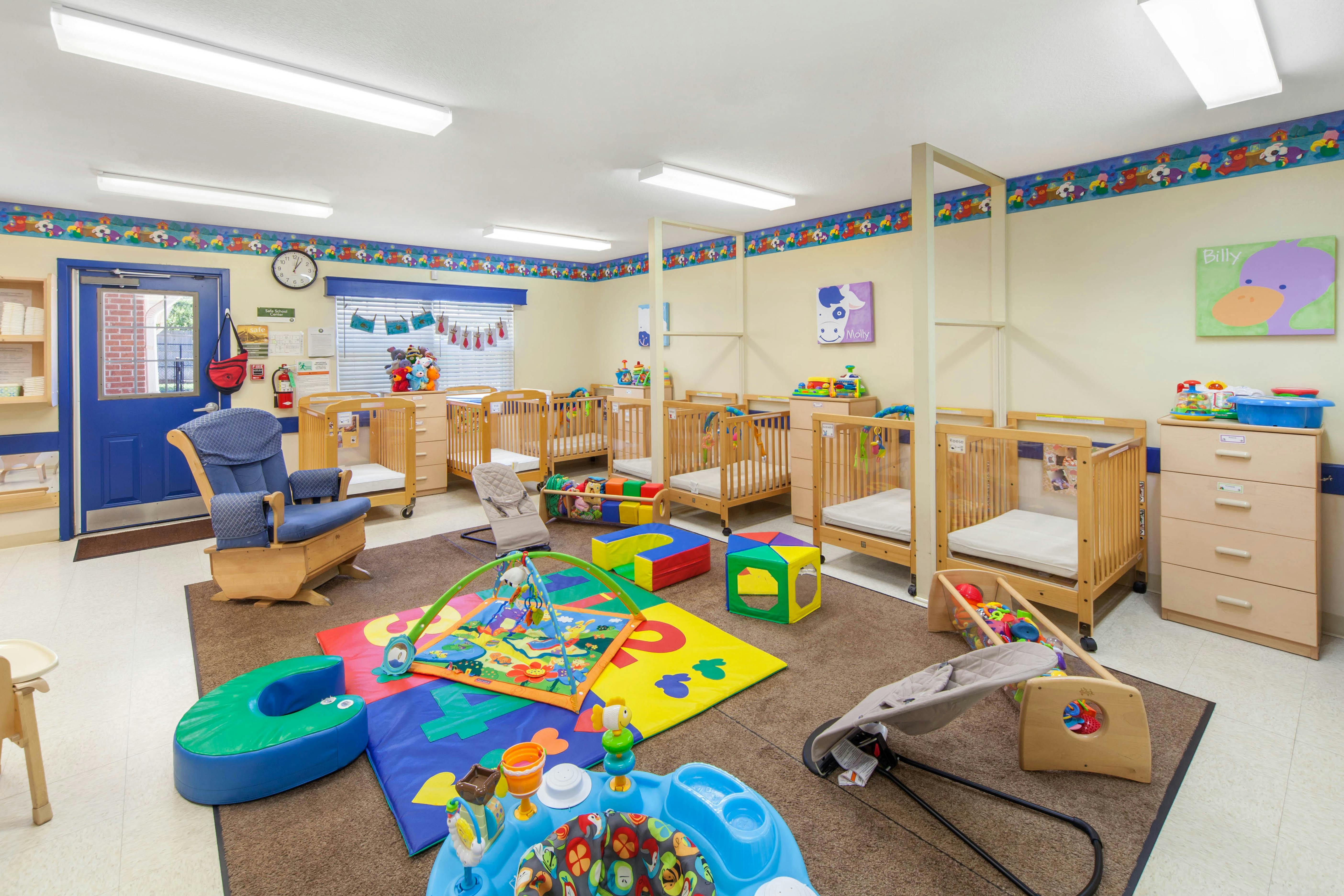 A colorful childcare room with cribs, playmats, a rocking chair, and a blue door