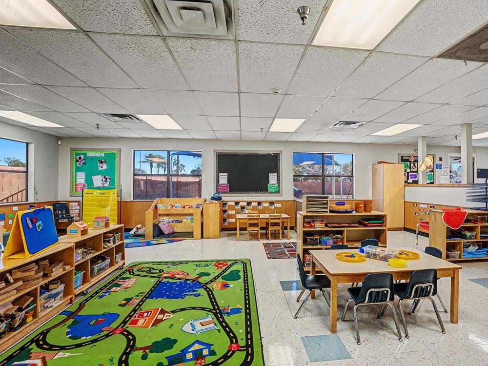 Classroom with child-sized tables and chairs, shelves with toys, and a road map rug