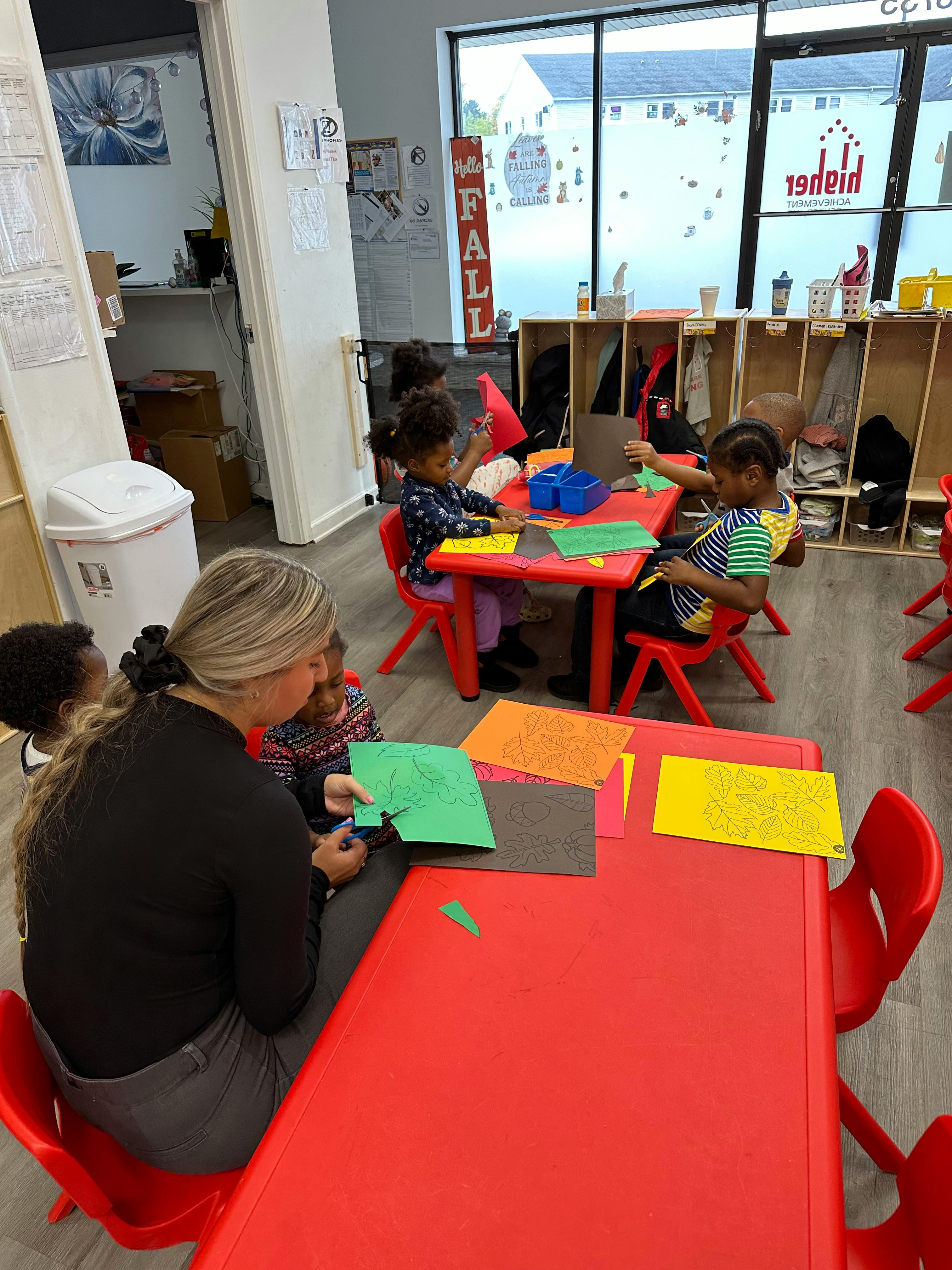 Adult and children cutting paper at red tables in a classroom