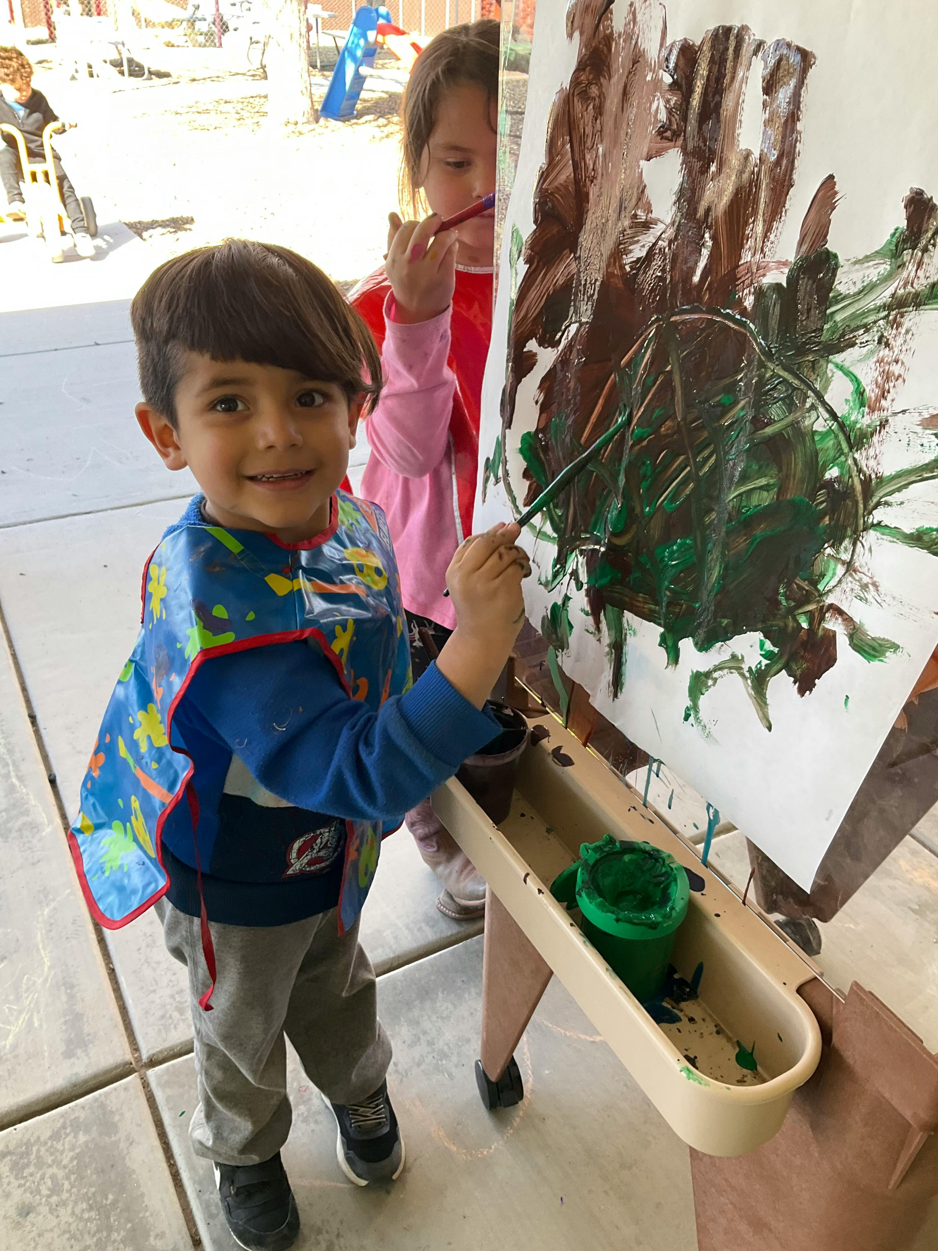 Child in a smock painting on an easel with another child in the background