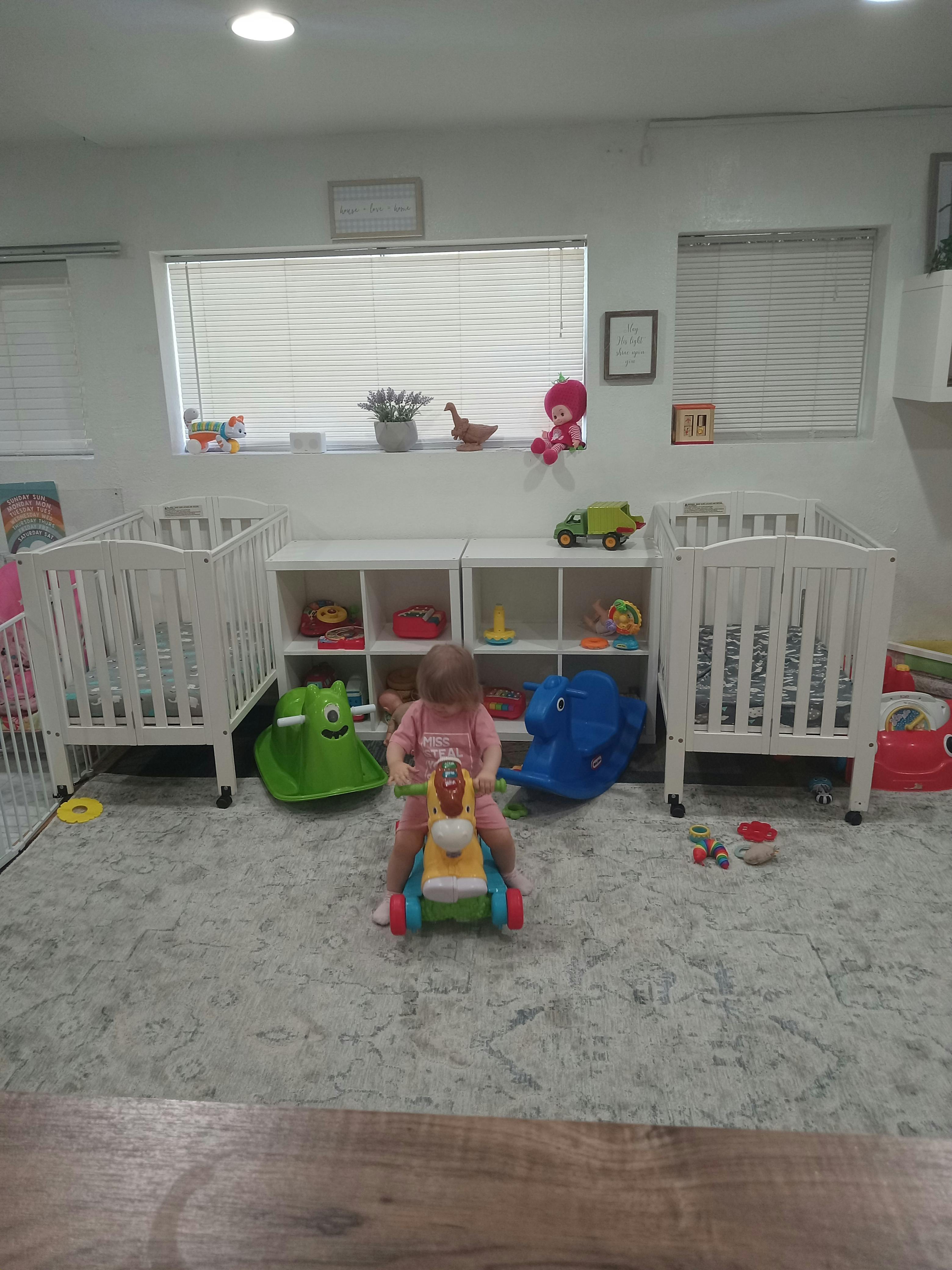 Child riding a toy horse in a room with cribs and shelves of toys
