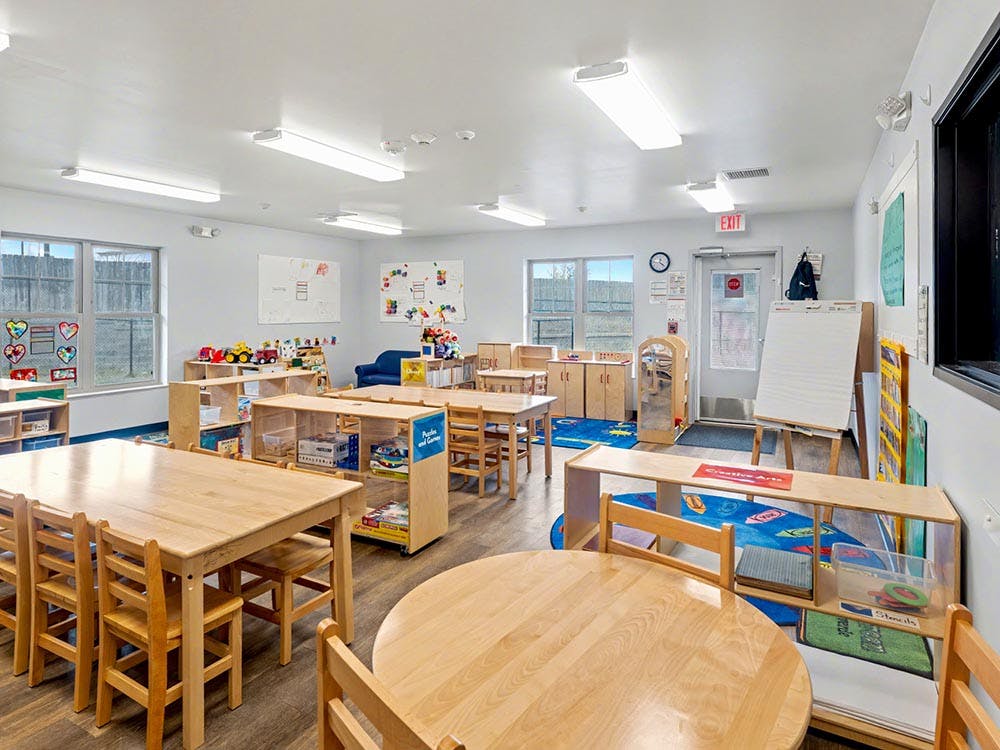 Classroom with wooden tables and chairs, shelves with toys, and whiteboard