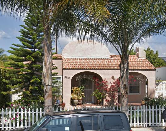 House with arched entryway and red tile roof behind a white picket fence