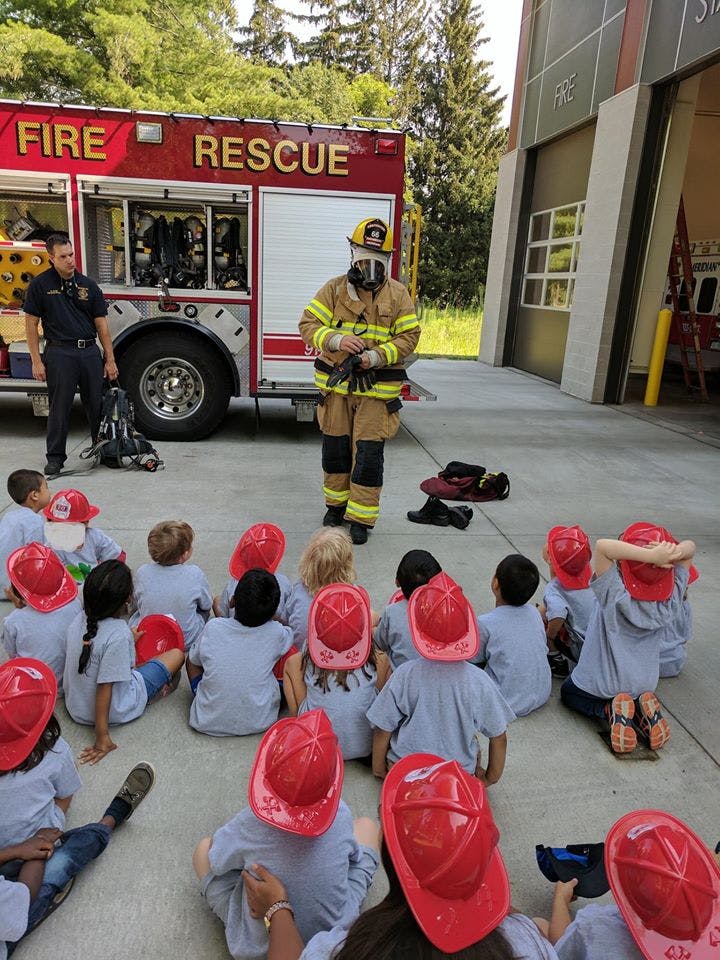 Firefighter in gear talking to children in red hats sitting on the ground