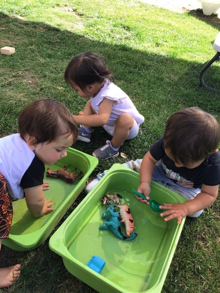 Three children playing with water and toys in green bins on grass