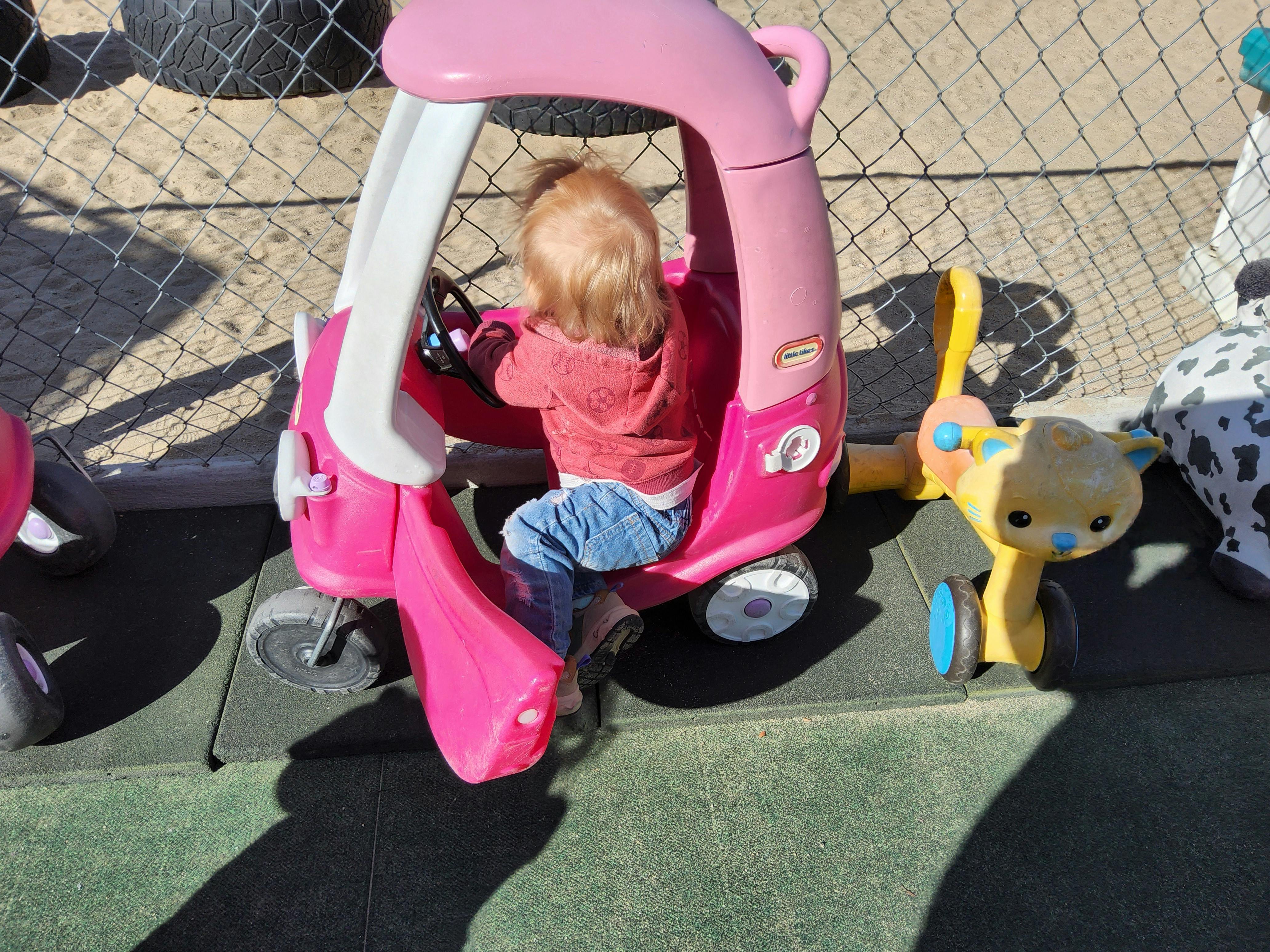Child lying upside down in a pink toy car with yellow handles visible next to it on an outdoor surface