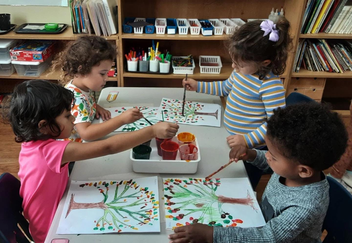 Four children painting tree pictures at a table in a classroom