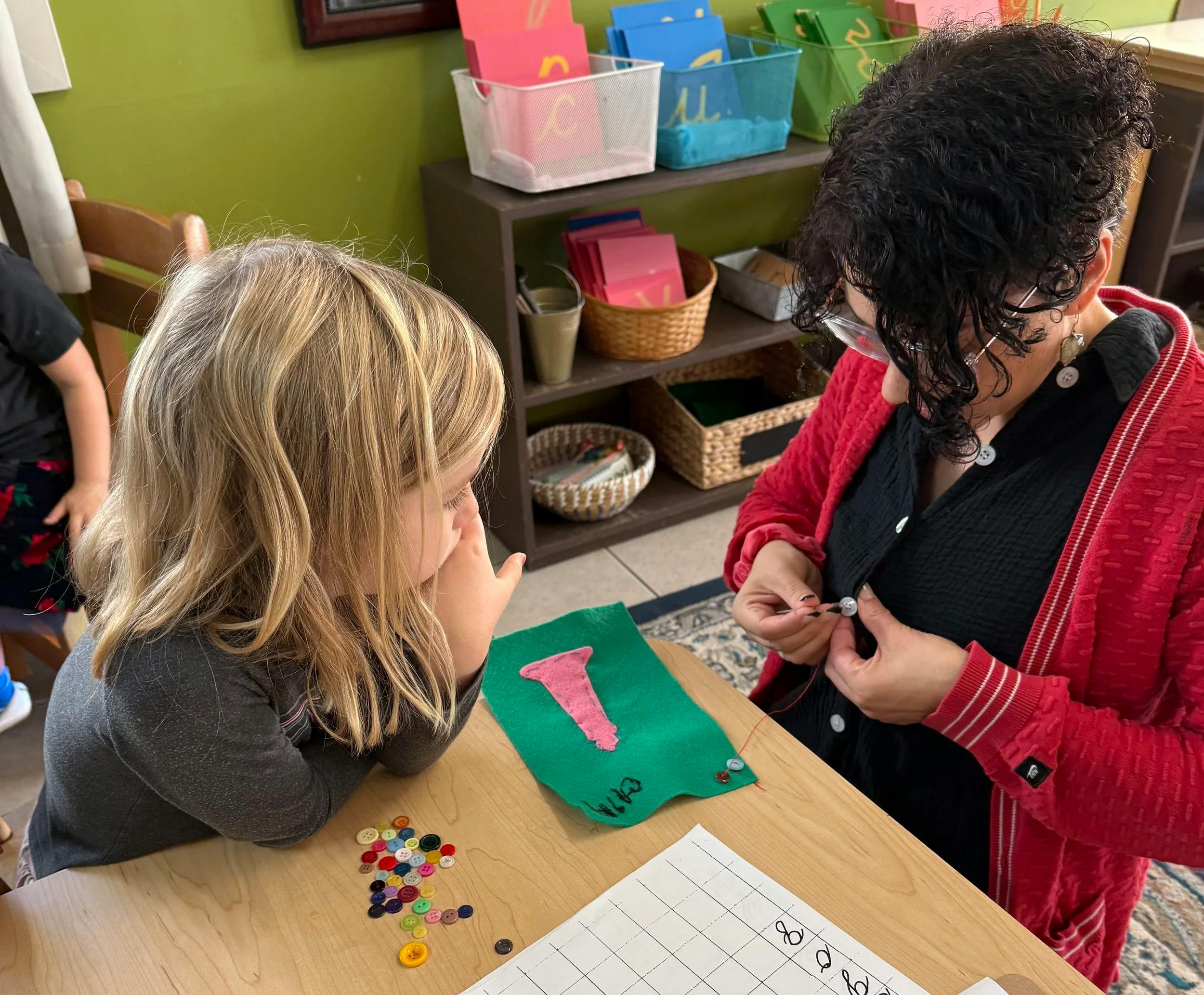 Adult and child learning to sew buttons onto fabric in a classroom