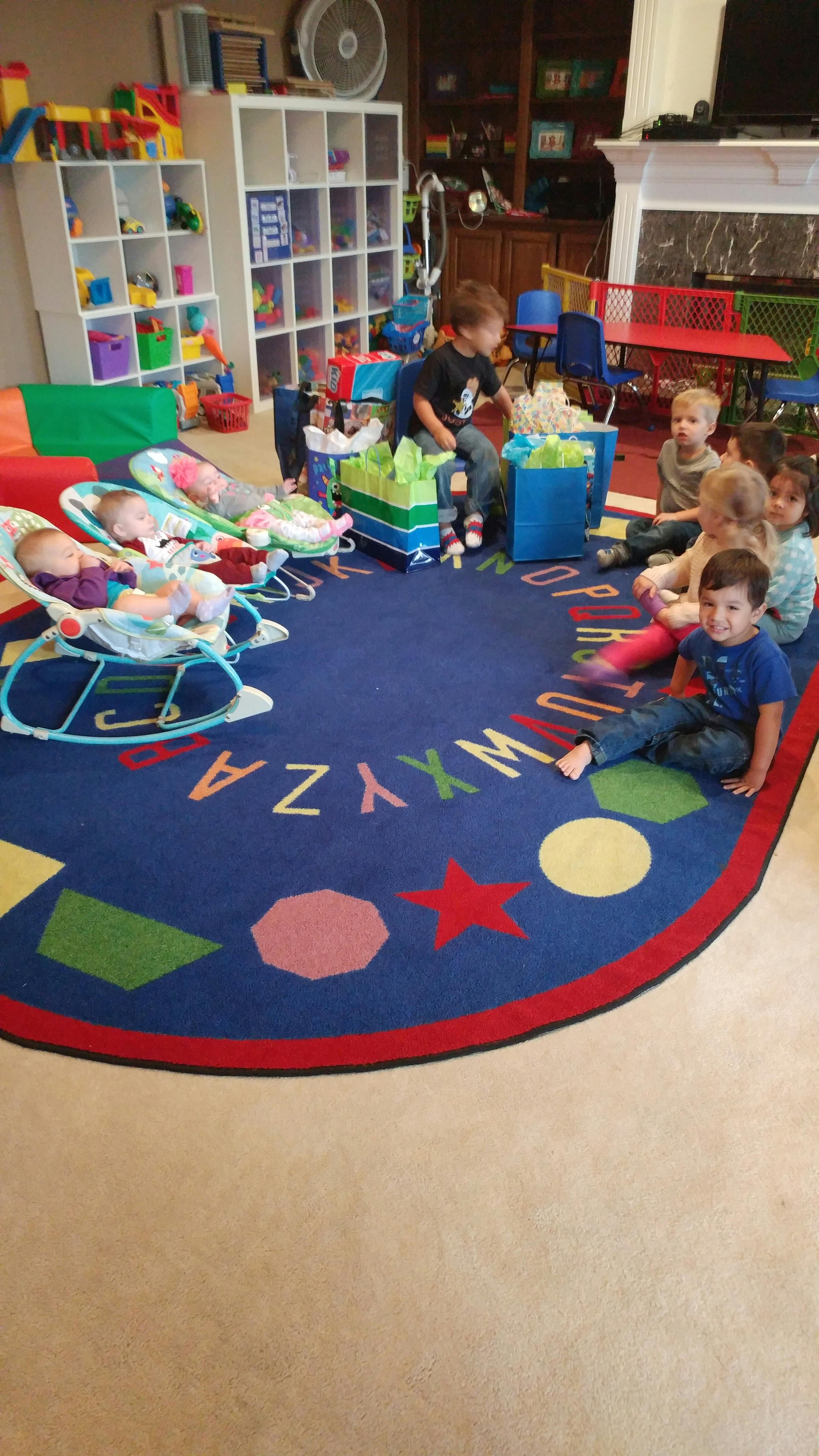 Several children gathered around gifts on a colorful alphabet rug, with babies in bouncers nearby