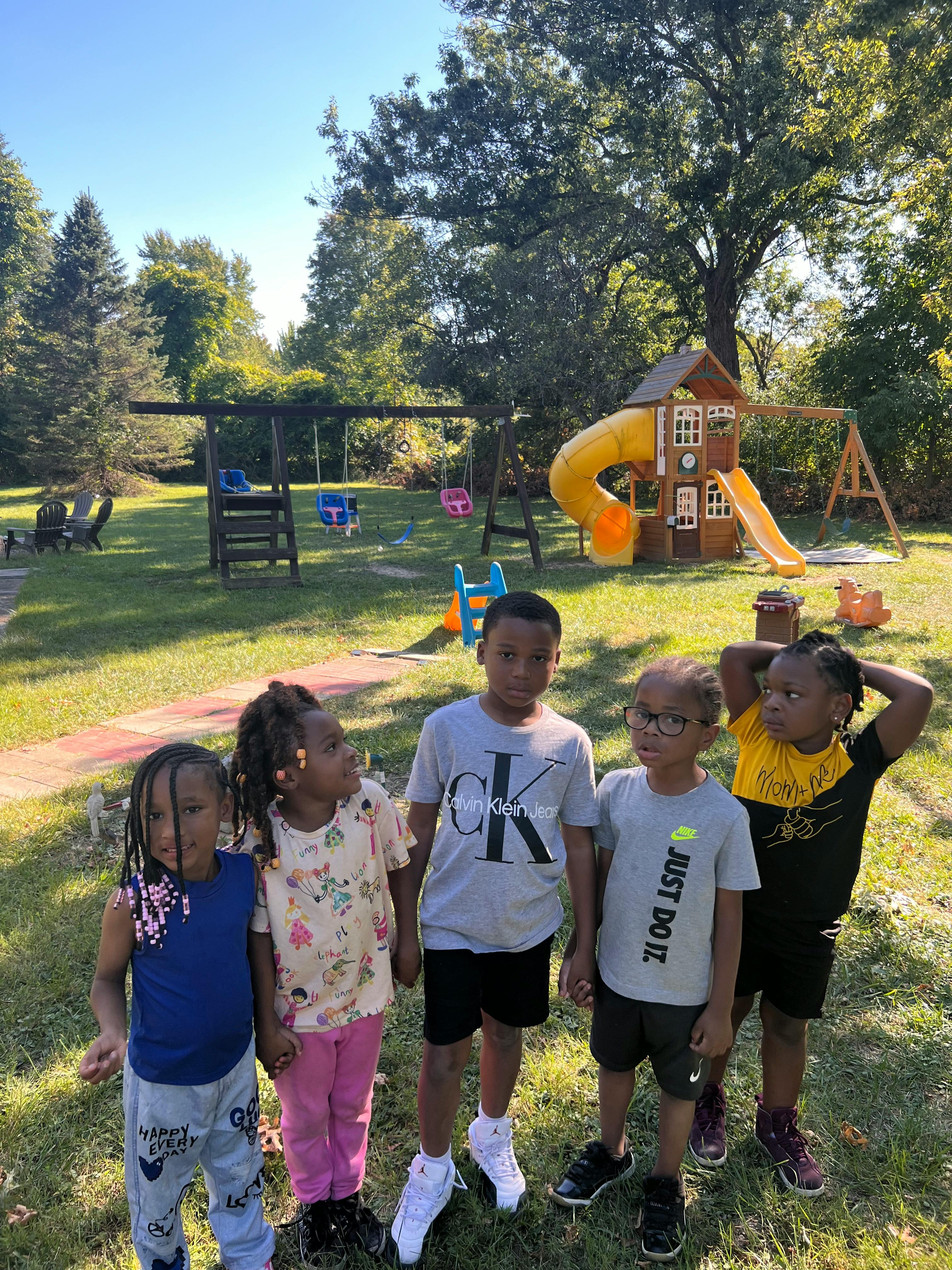 Five children standing in a grassy area with a playground in the background