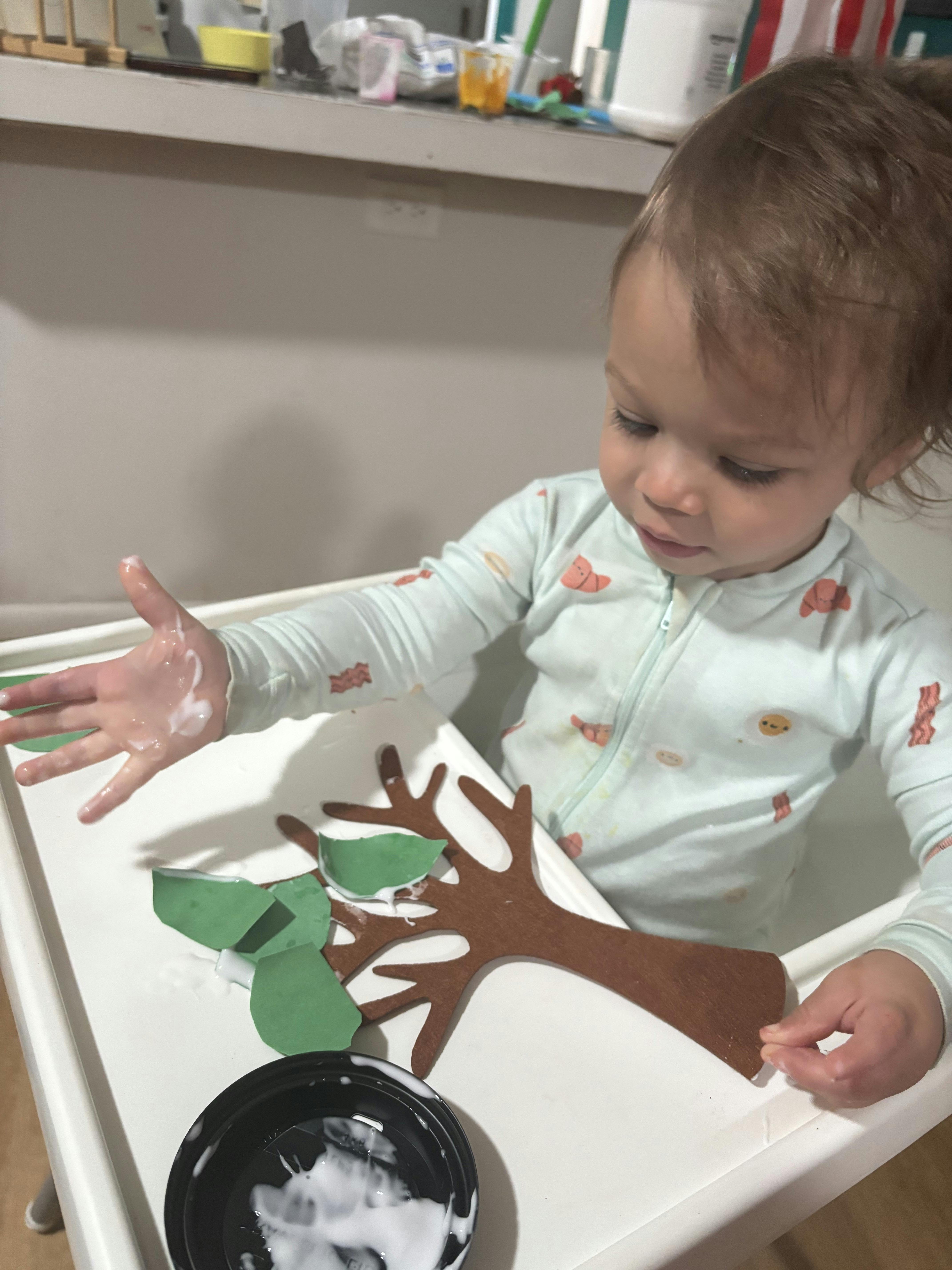 Child crafting with paper leaves and a wooden tree cutout on a high chair