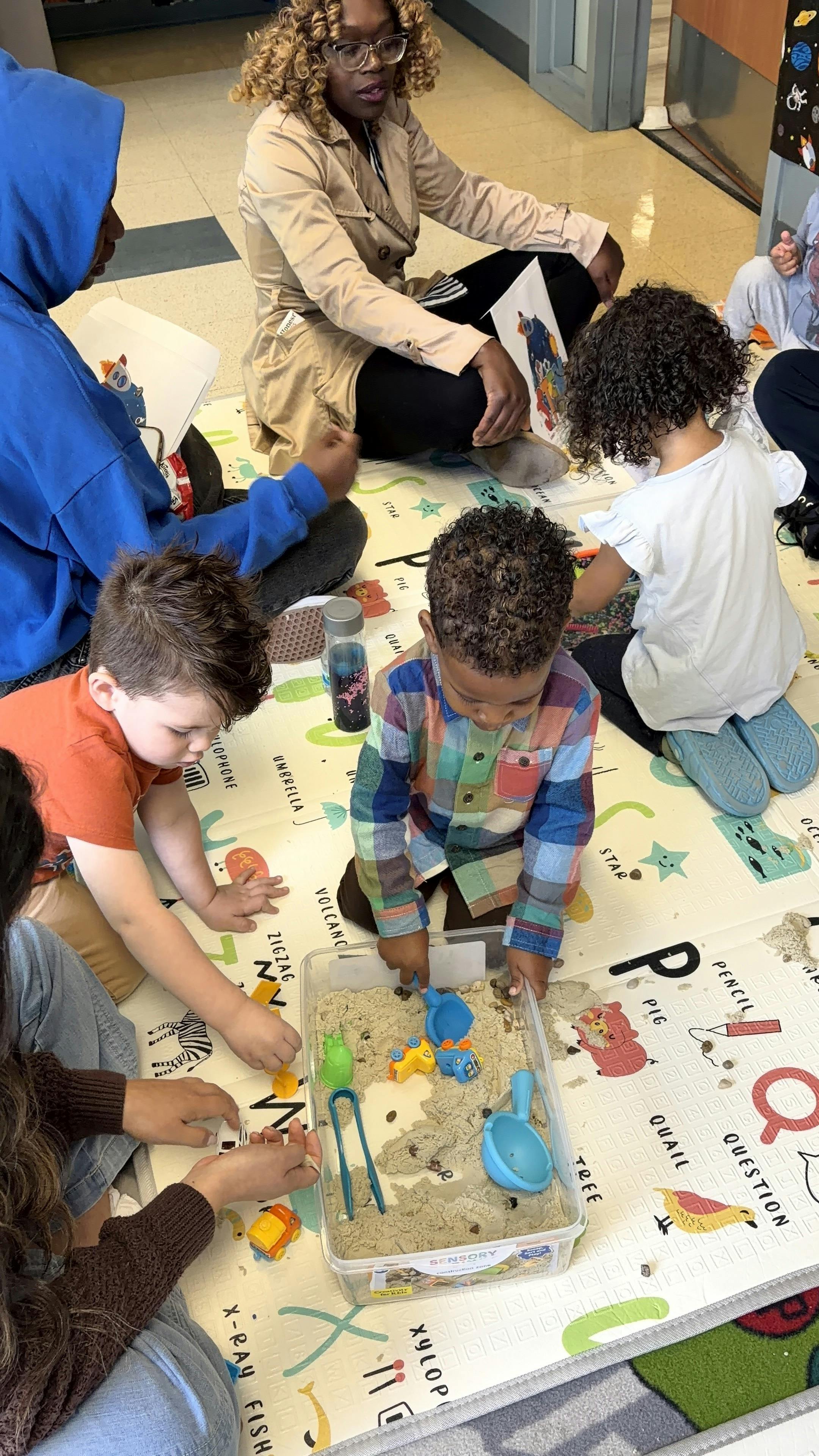 Adults and children on a mat, some playing with sand in a bin, others holding books