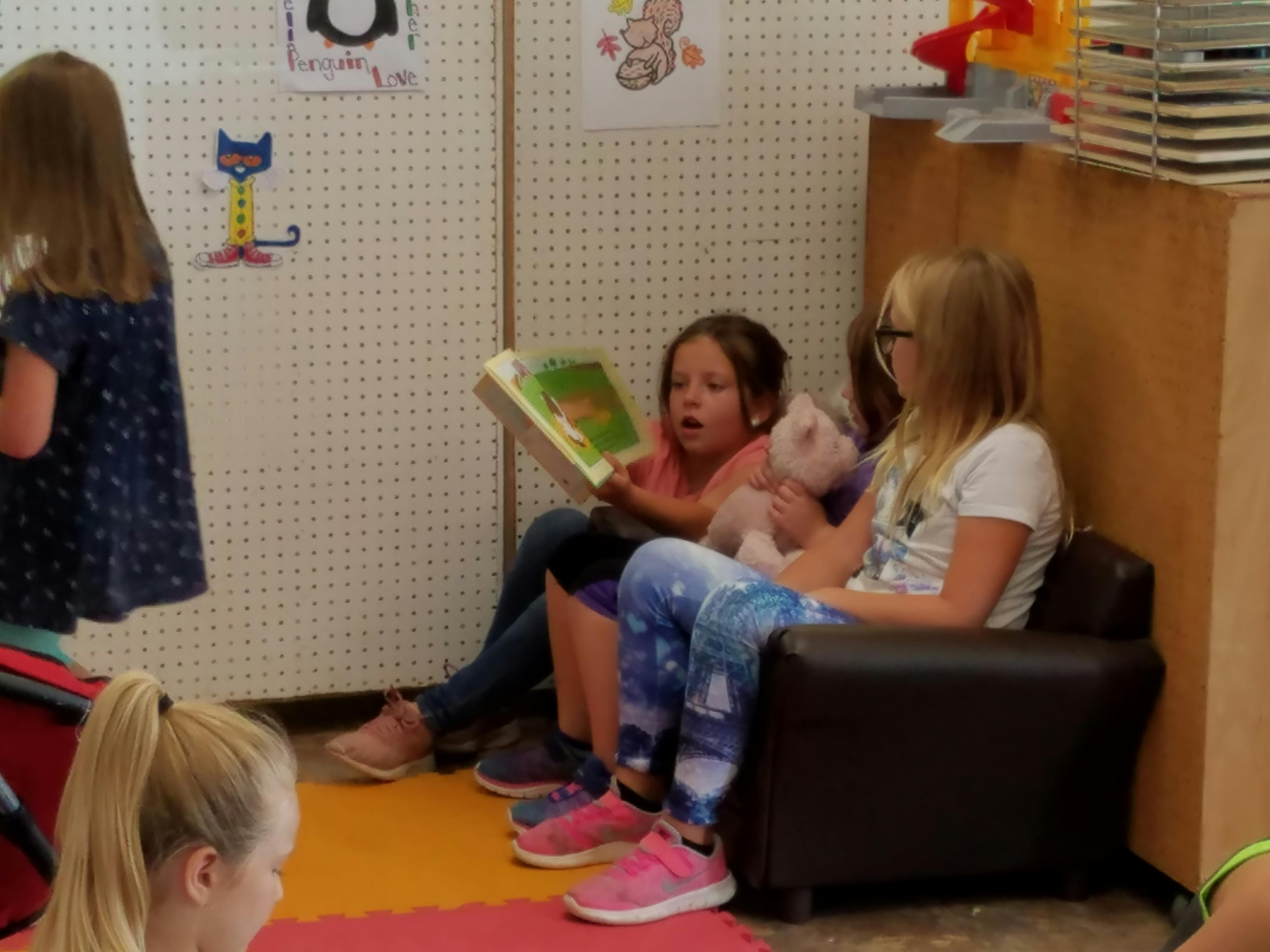 Two children sitting in a chair, one reading a book aloud