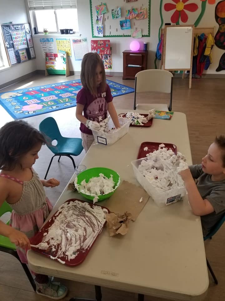 Three children playing with shaving cream at a table in a classroom