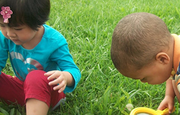 Two children playing in grass, one looking through a magnifying glass