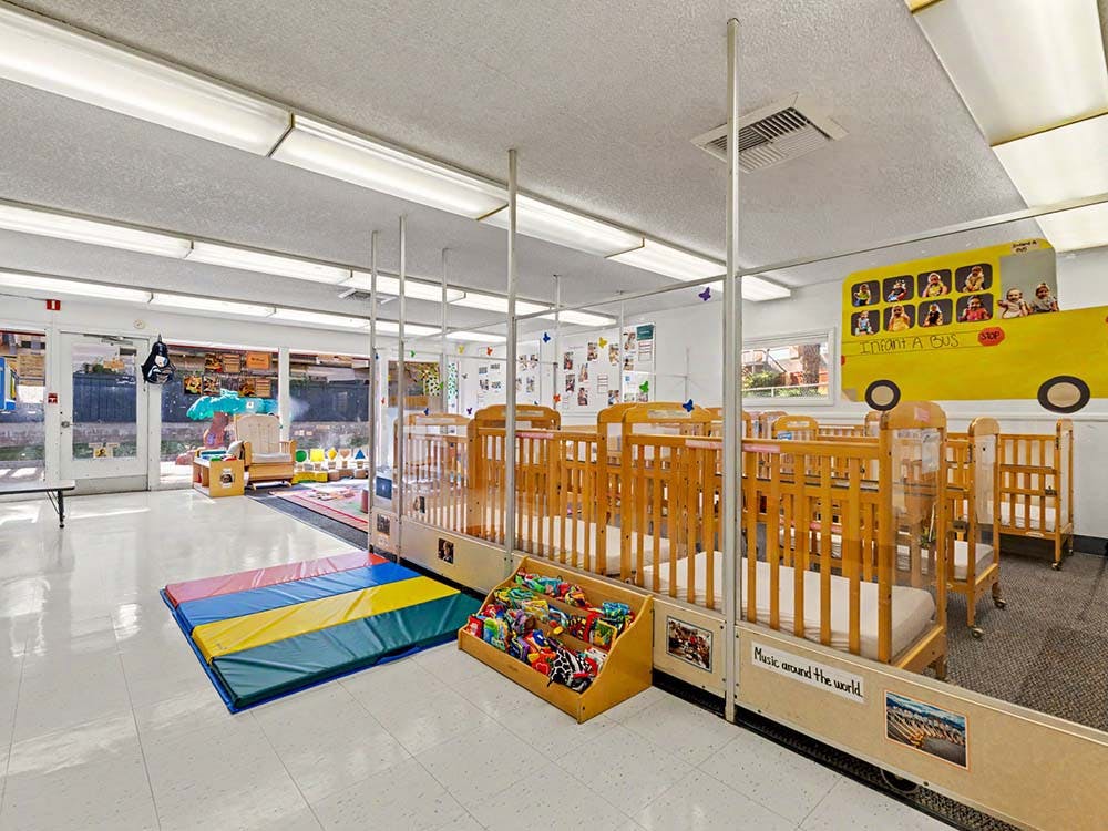 cribs in a daycare room with a colorful play mat and school bus decoration