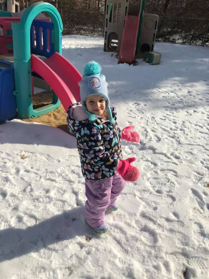 Child in winter clothing standing in snow next to a playground slide